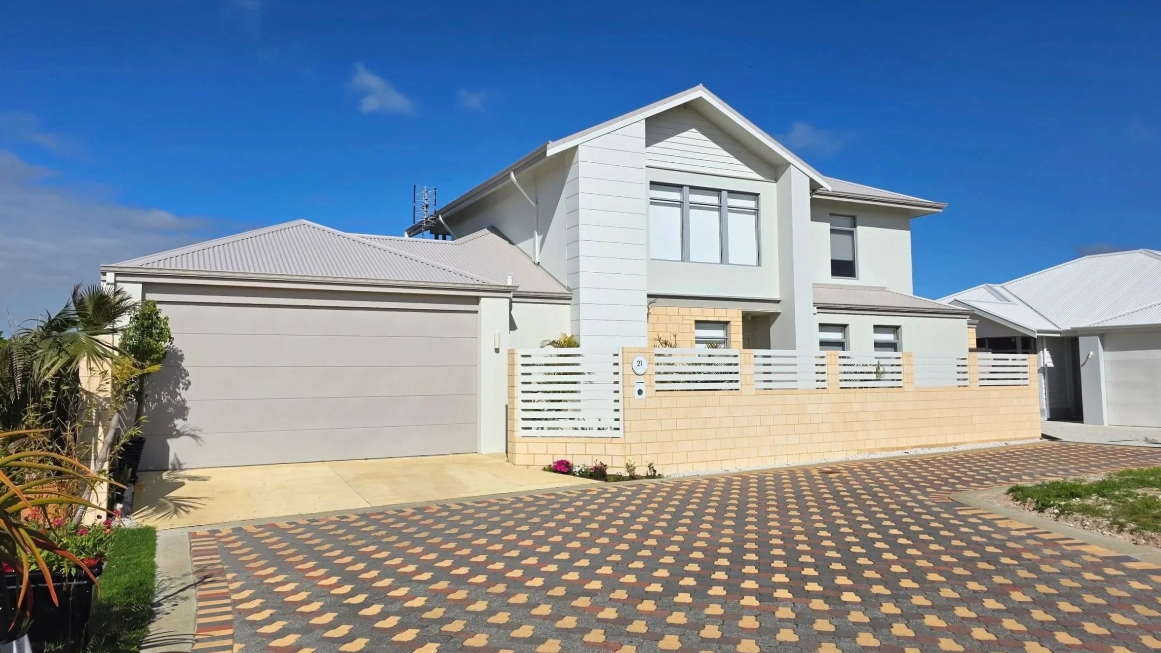 Modern two-story house with white exterior walls and a gray roof, surrounded by a fenced front yard and paved driveway, under a bright blue sky.