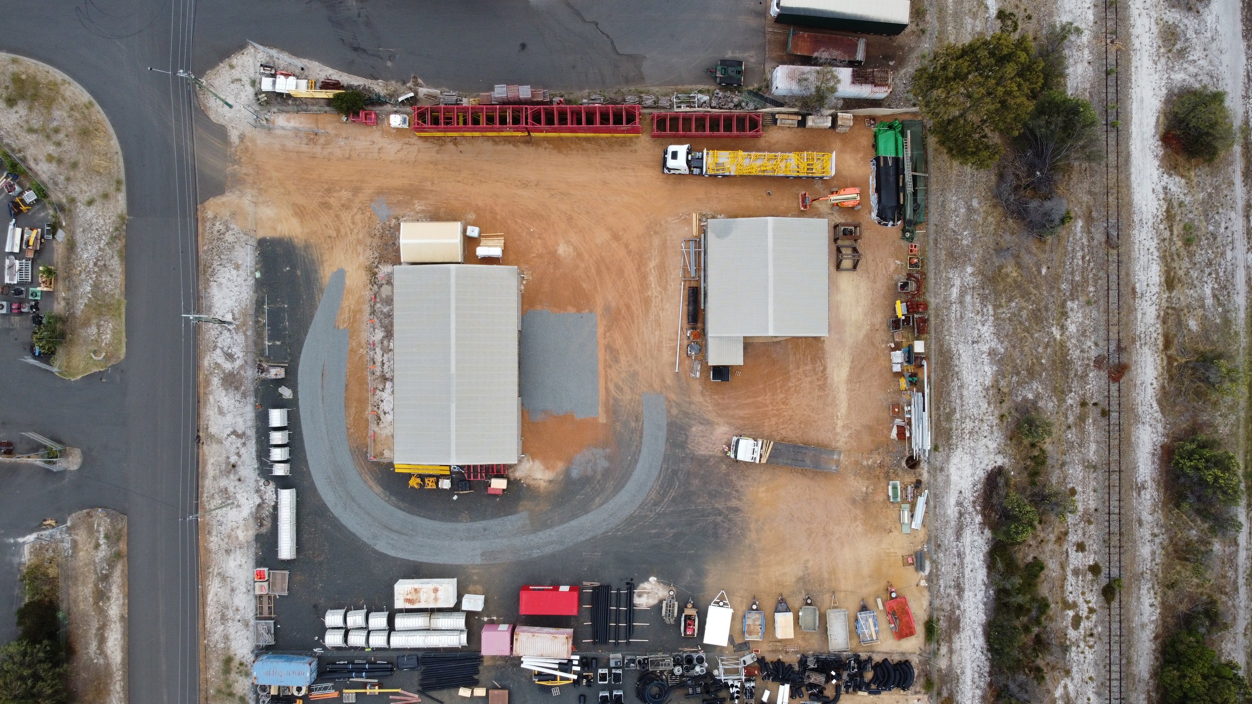 An aerial view of a construction site with two buildings, construction equipment, materials, and a dirt and paved area surrounded by trees and railway tracks.