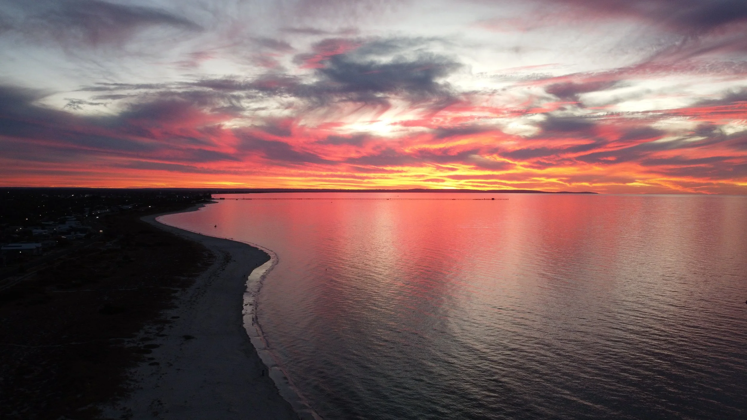 A scenic view of a beach at sunset with colorful clouds in the sky and calm water reflecting the sunset hues.