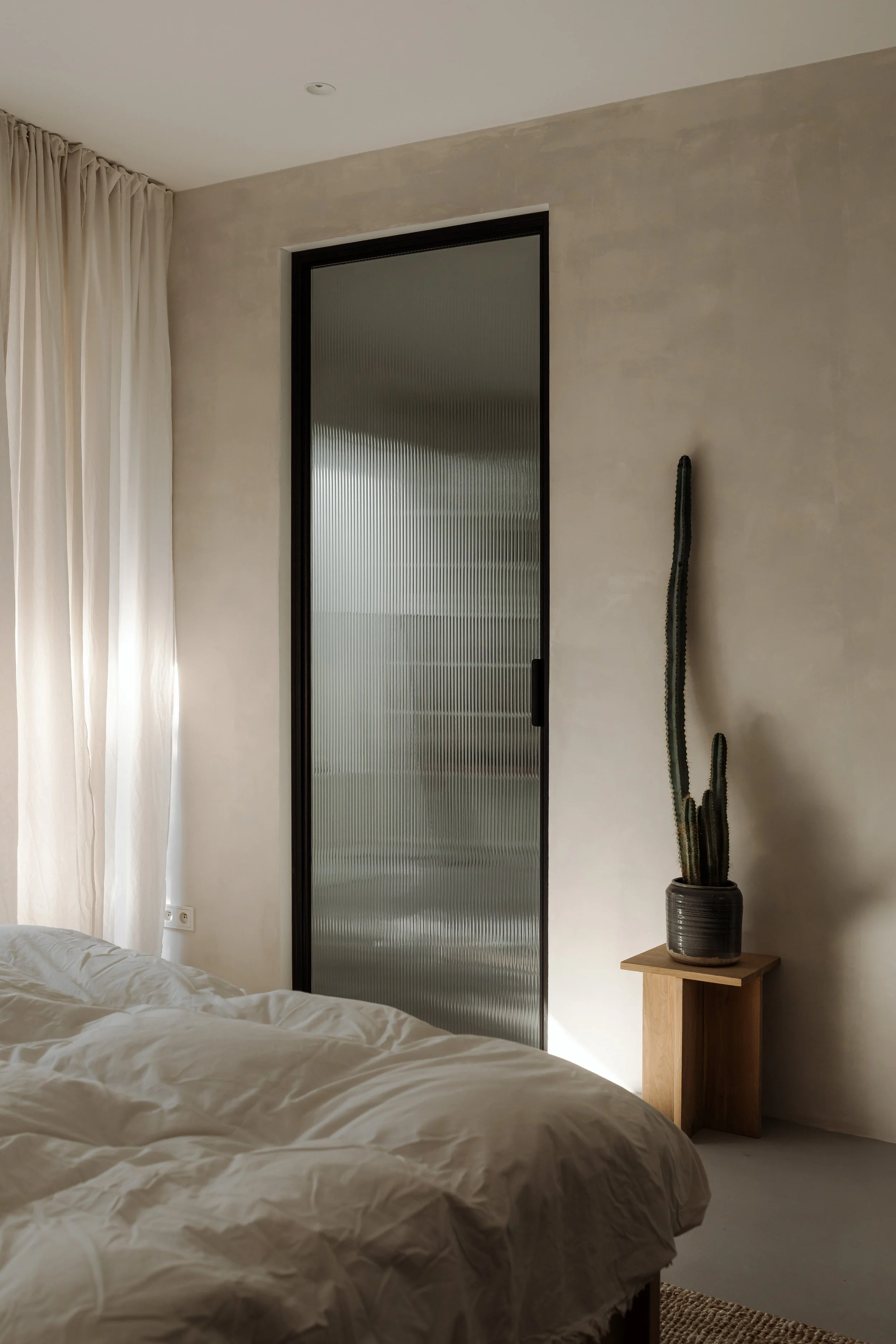 A bedroom with a bed, beige curtains, a frosted glass door, and a tall cactus plant in a black pot on a wooden stand.
