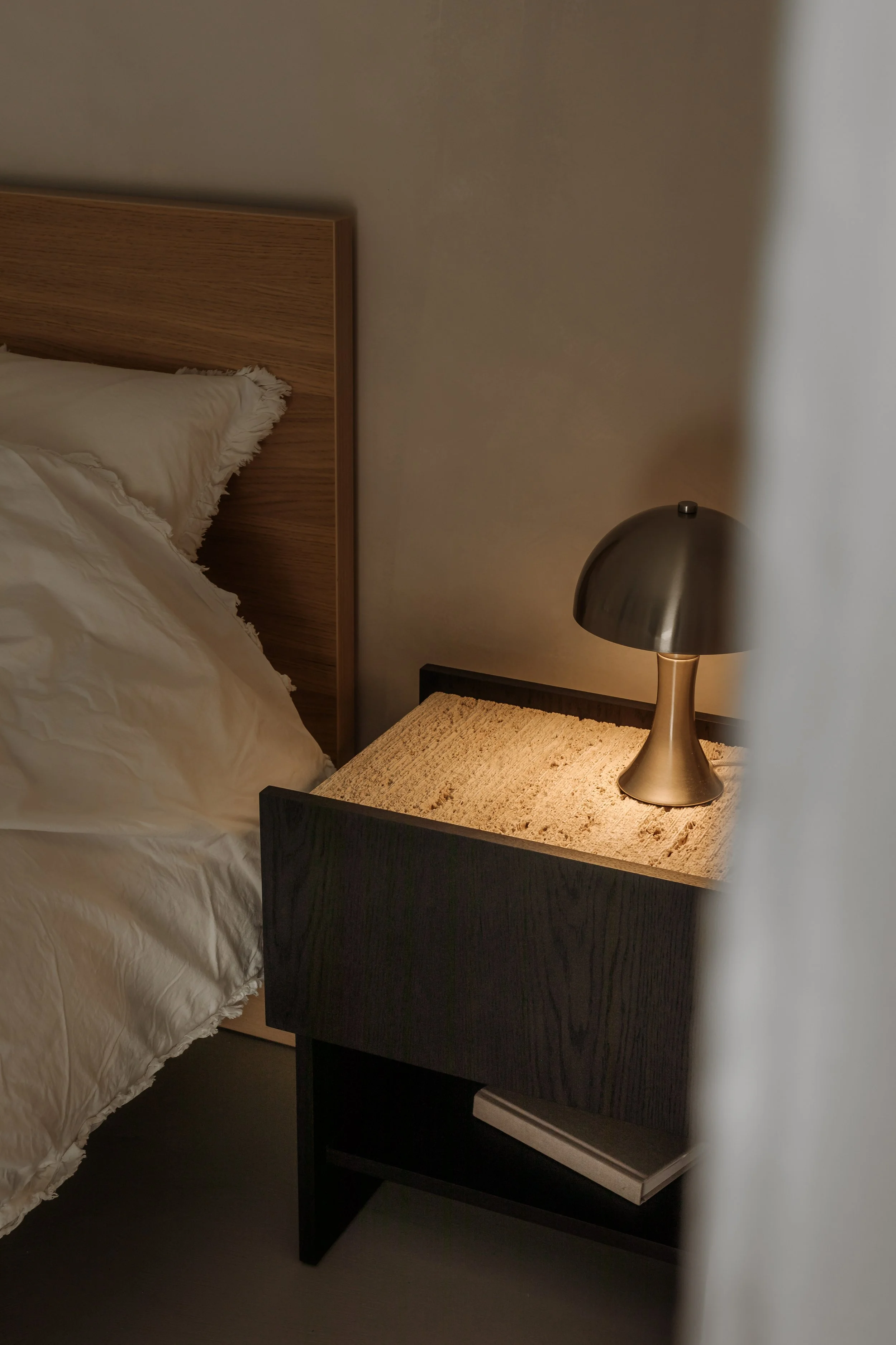 A bedroom corner with a wooden bed headboard, white rumpled bed sheets, and a nightstand with a modern metallic lamp and a book underneath.