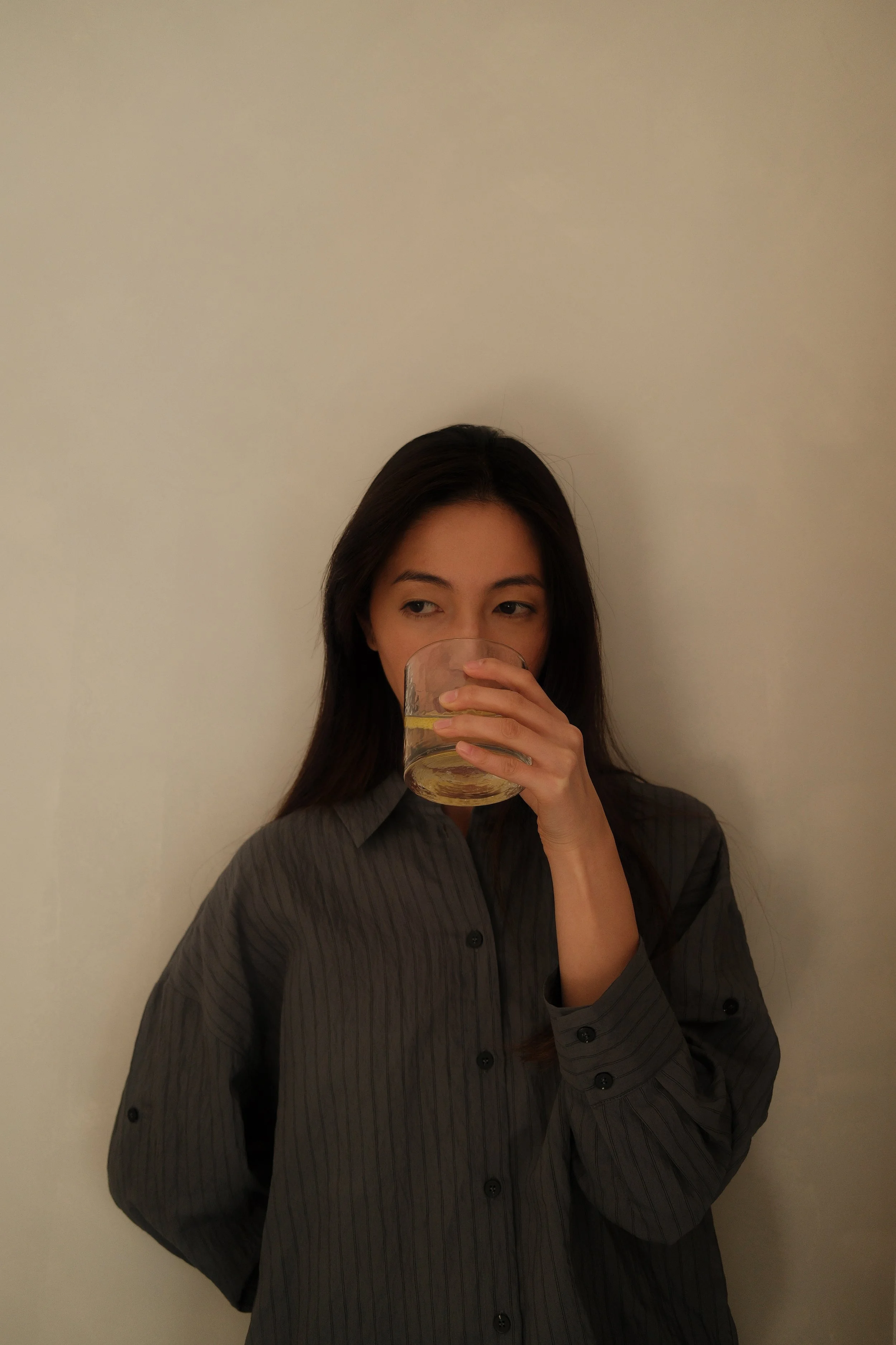 A woman with long dark hair, wearing a dark button-up shirt, is standing against a plain light-colored wall, drinking from a glass of water with a lemon slice inside.