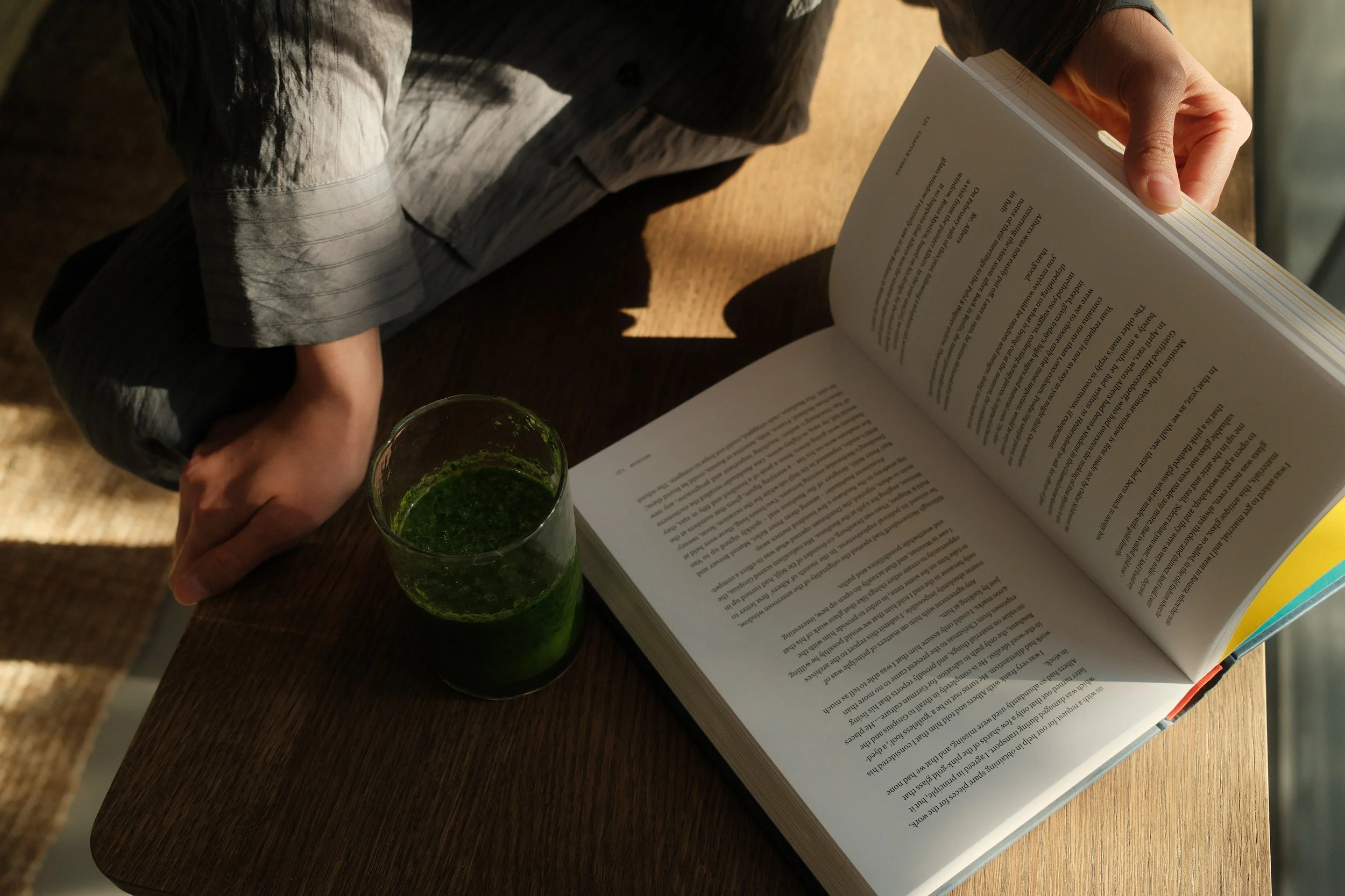 Person sitting at a wooden table reading a book with a glass of green juice nearby.