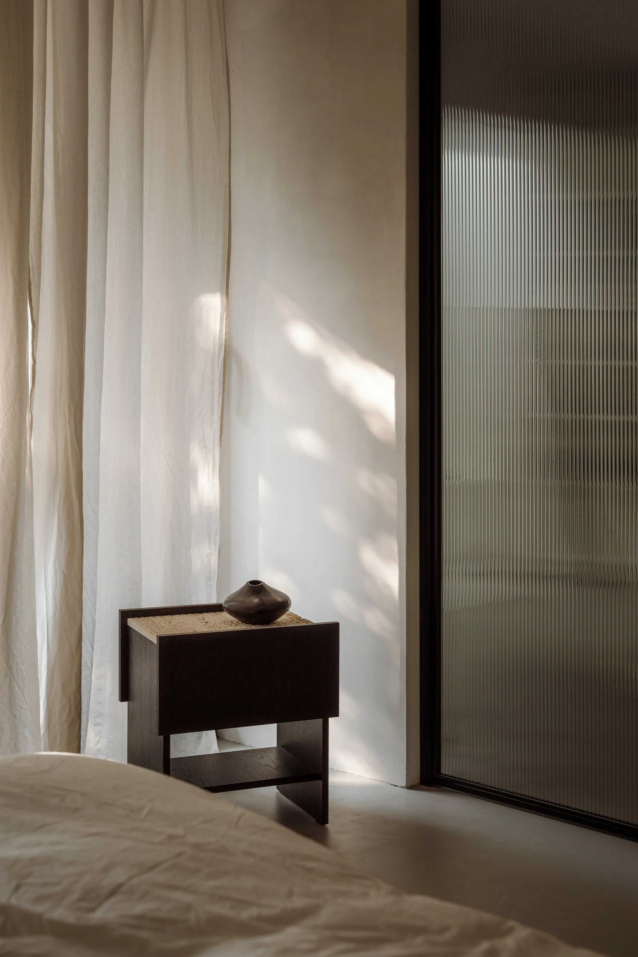 Interior bedroom scene with a bed in the foreground, a small dark wooden bedside table with a decorative vase on top, white curtains, and a textured glass door.