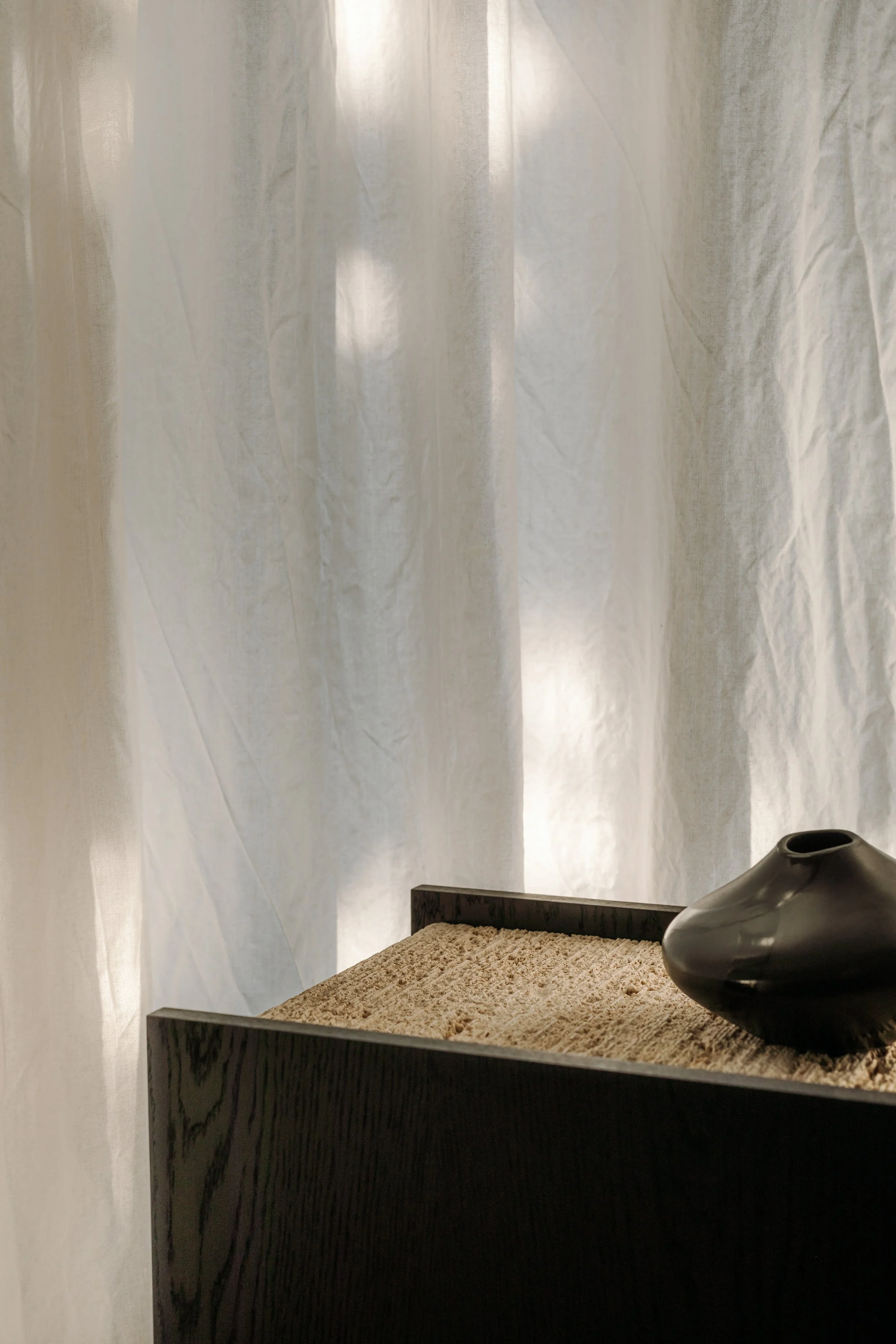 A black vase on a wooden surface with beige sand, set against white curtains.
