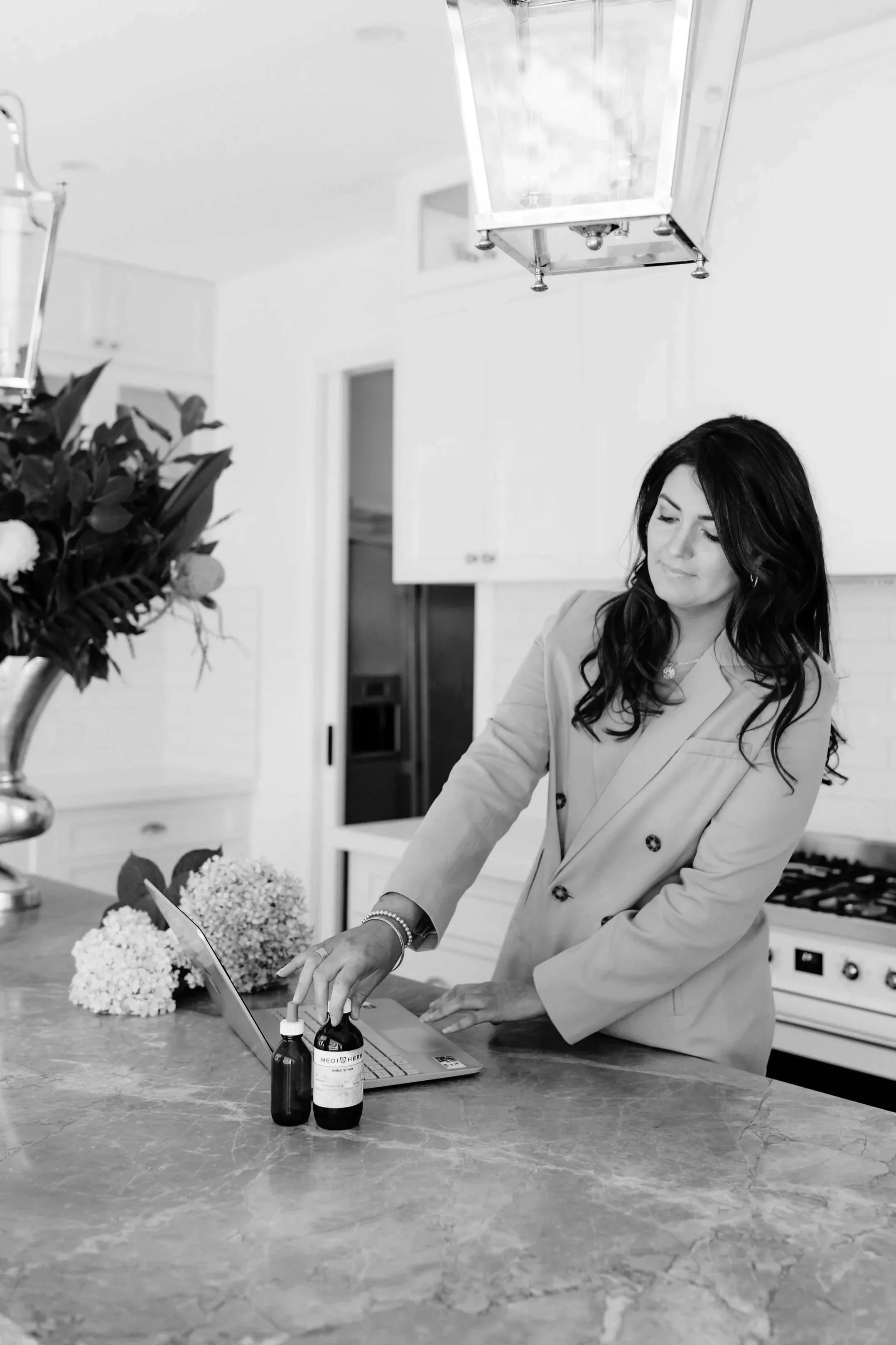 A woman with dark hair and wearing a light-colored blazer is in a kitchen, standing at a marble countertop. She is looking at some small bottles, possibly medication, on the countertop while using a laptop. The kitchen background includes flowers, cabinets, and a stove.