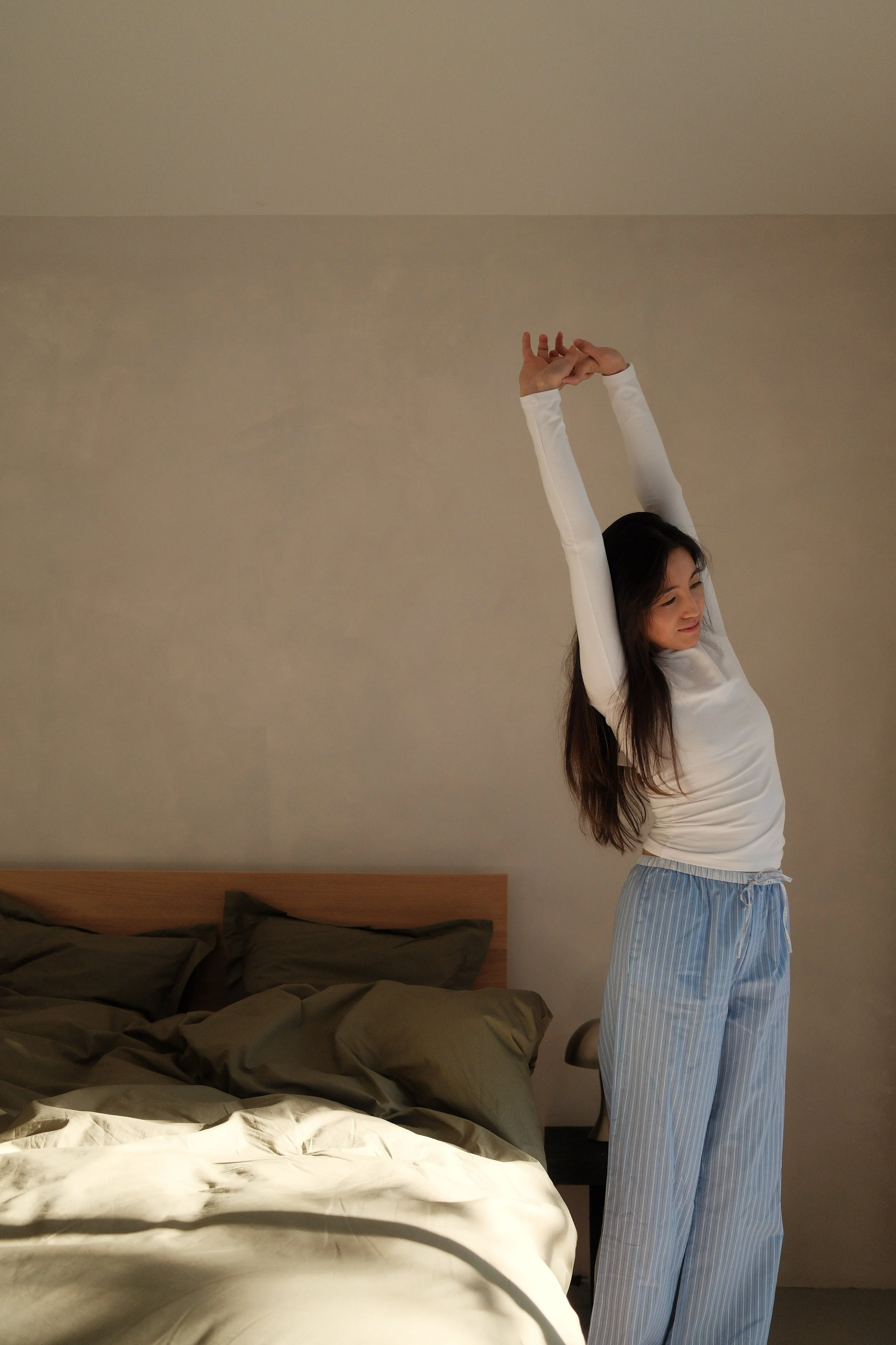 A woman stretching in pajamas next to a bed in a bedroom.