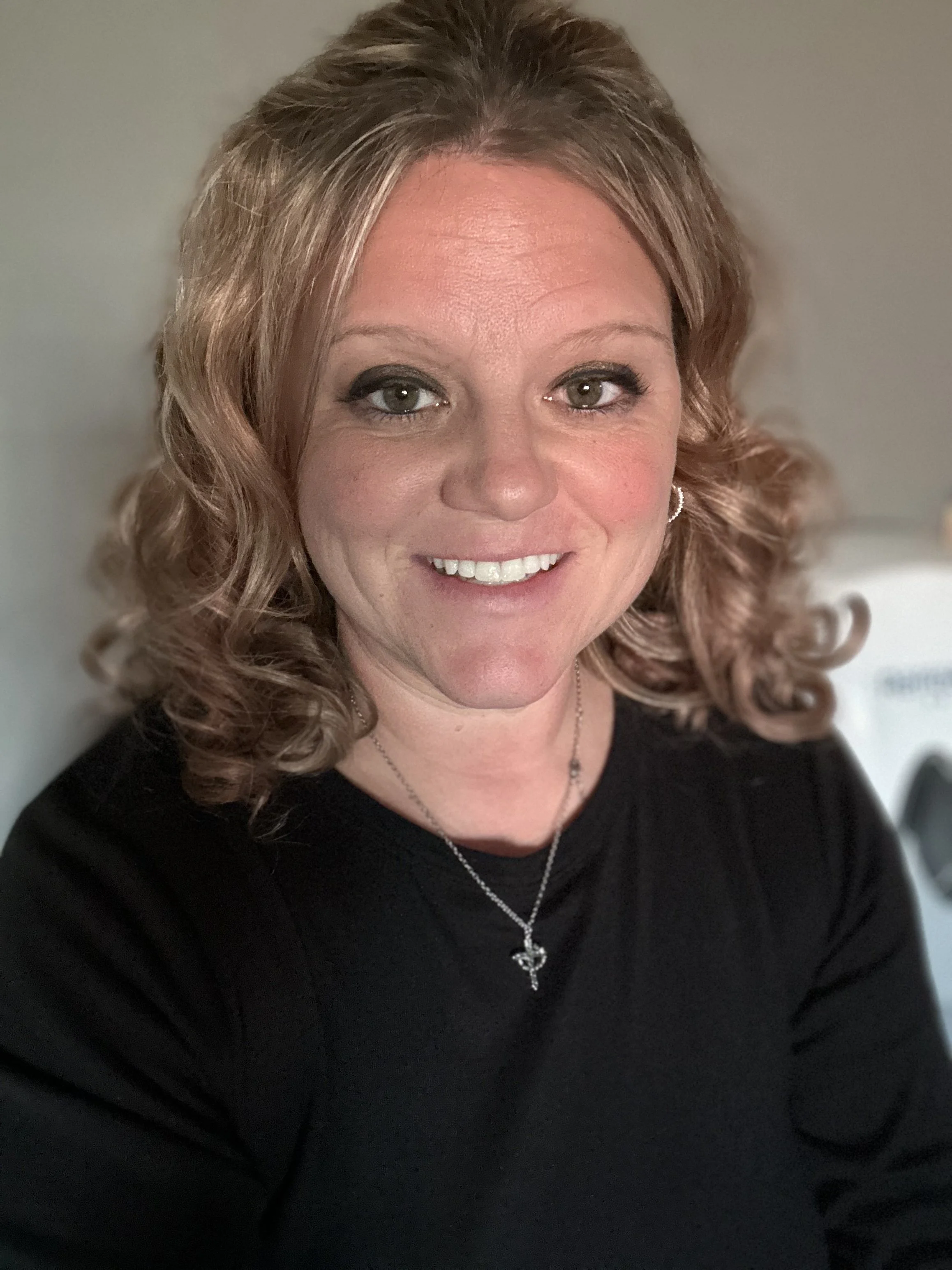 A woman with wavy, shoulder-length red hair, smiling, wearing a black top, a silver cross necklace, and earrings, against a neutral background.