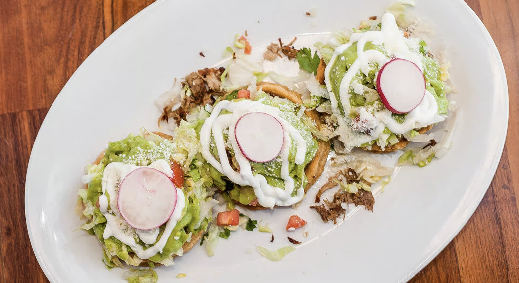 Three tacos on a white oval plate with shredded lettuce, radish slices, and white sauce on top, served on a wooden table.