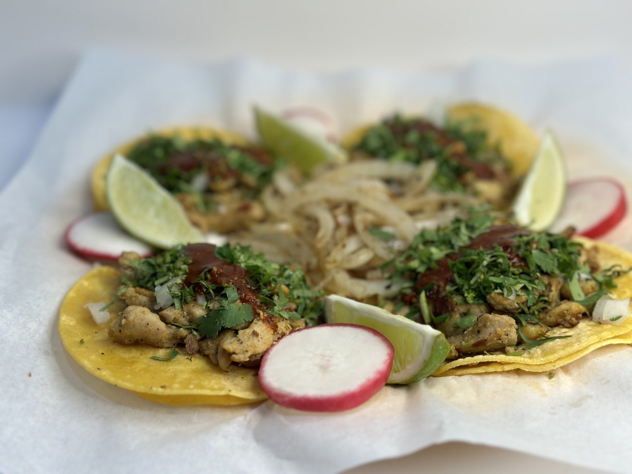Close-up of four chicken tacos, each topped with chopped onion, cilantro and sauce, with lime wedges and sliced radishes.