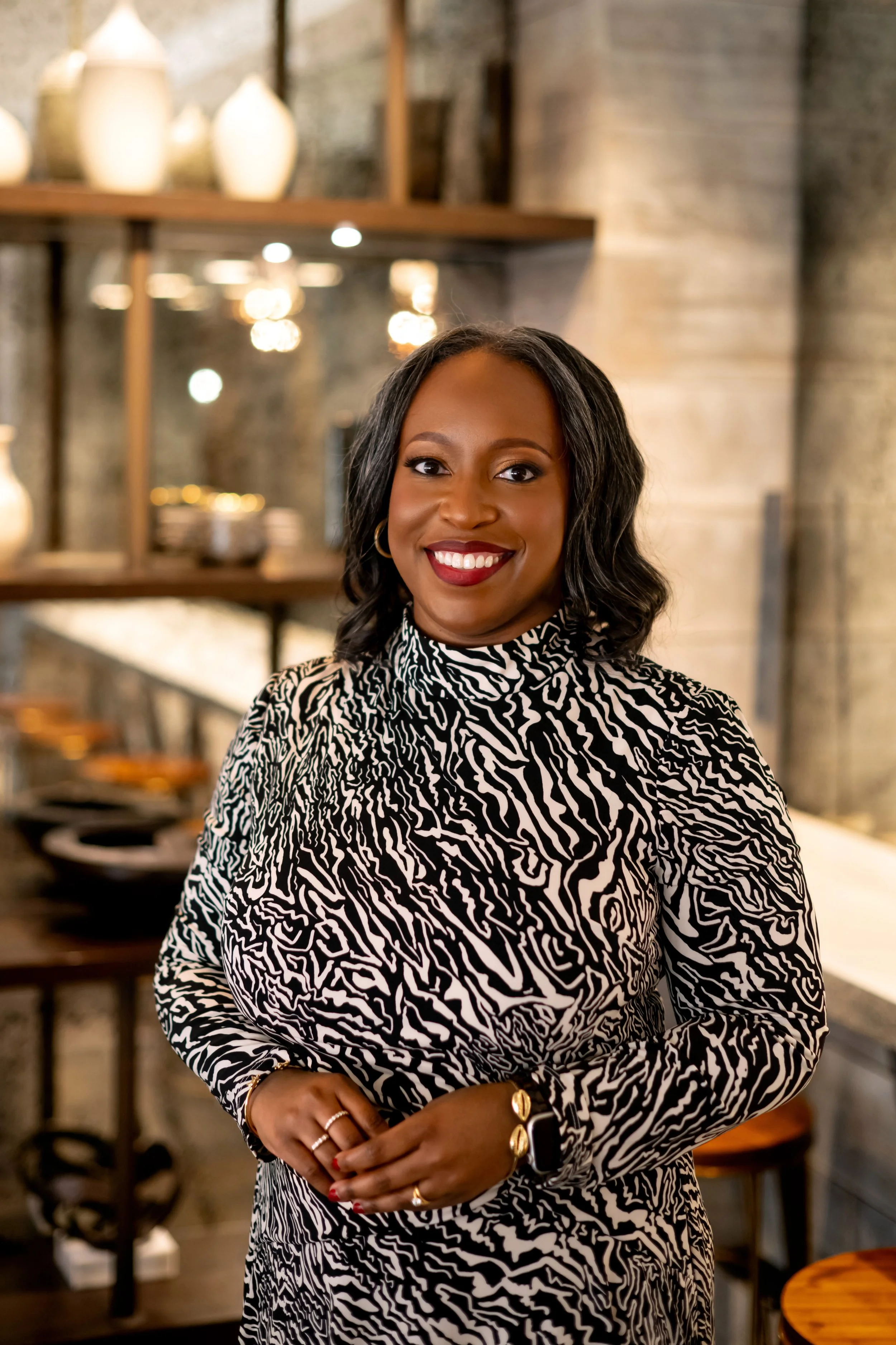 A woman with dark hair styled in loose waves, wearing a black and white animal print top, smiling, standing in a modern interior with decorative pottery on wooden shelves and warm lighting.
