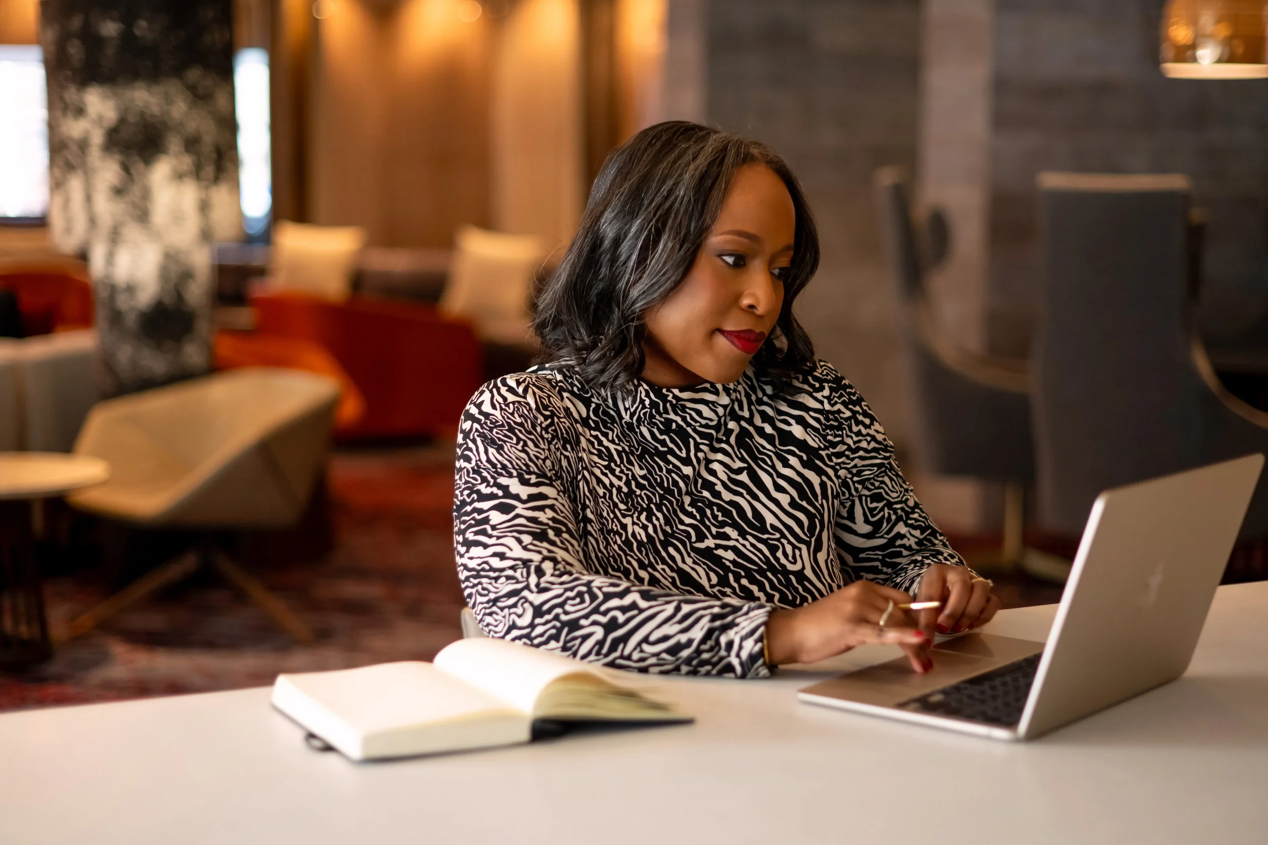 A woman with dark hair and red lipstick sitting at a table using a laptop in a cozy, modern lounge area with comfortable chairs and warm lighting.