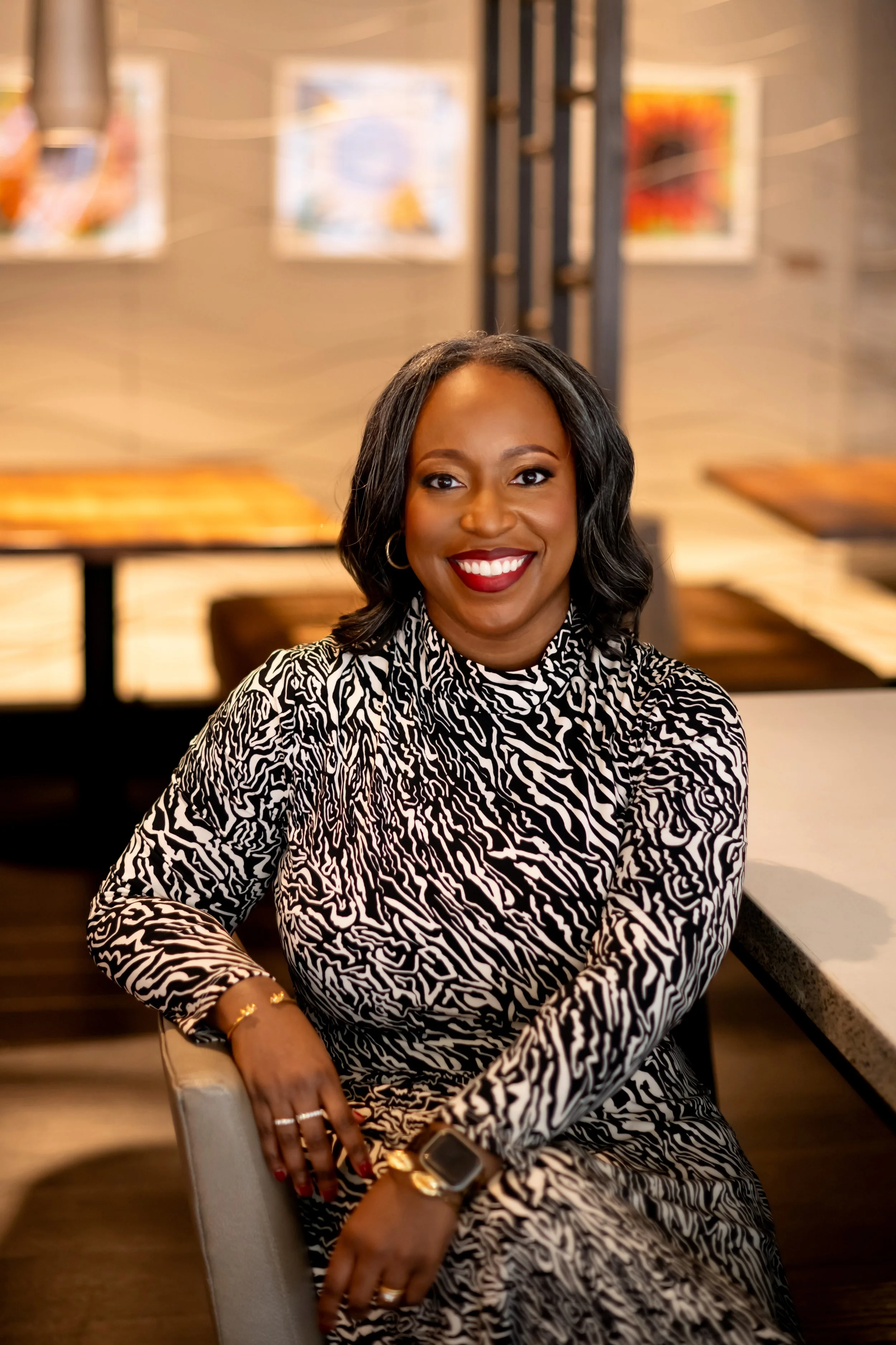 A woman with dark hair in a zebra-print dress sitting at a table, smiling in a well-lit restaurant or cafe.