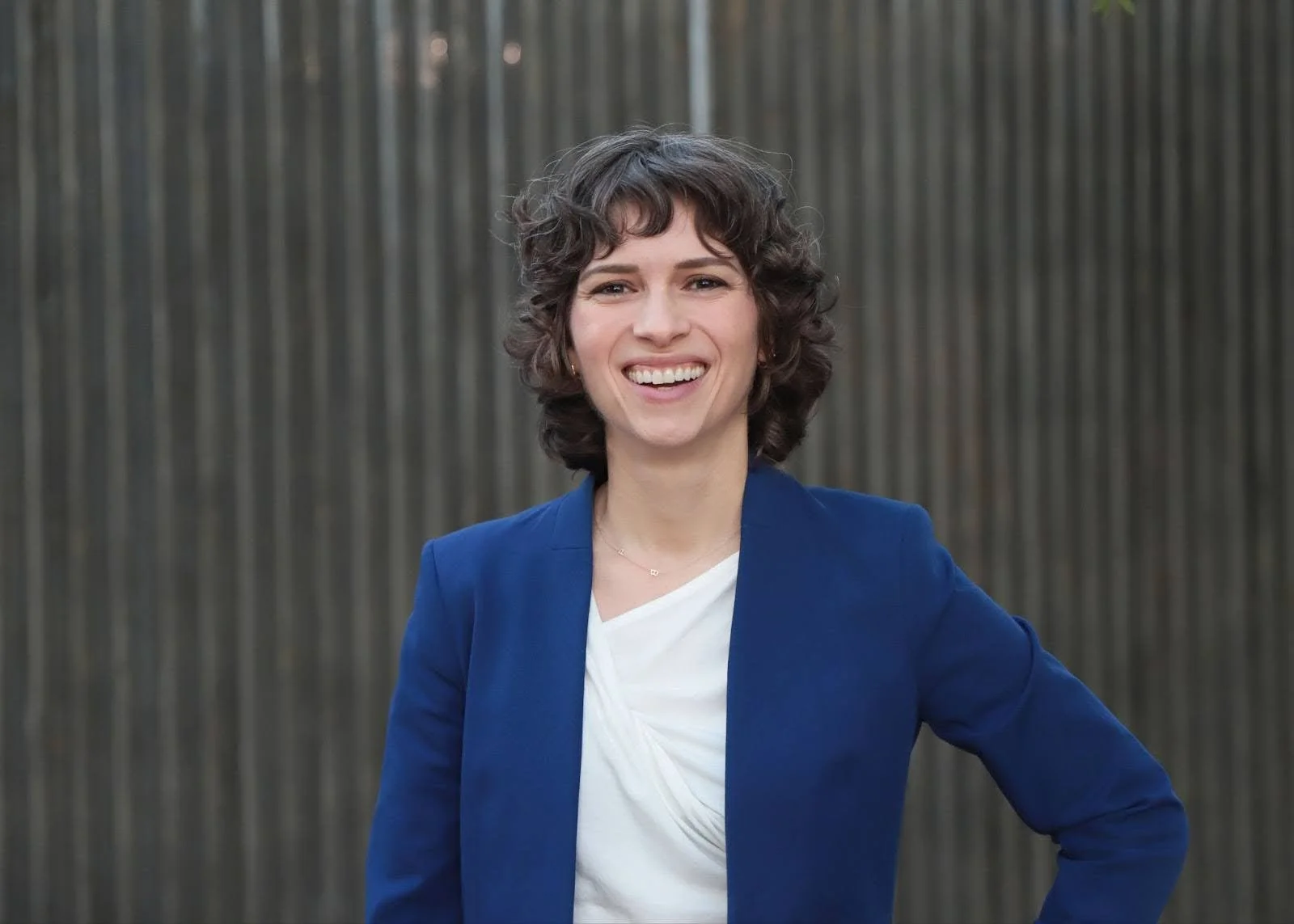 A smiling woman wearing a blue blazer and a white top, standing outdoors in front of a gray fence.