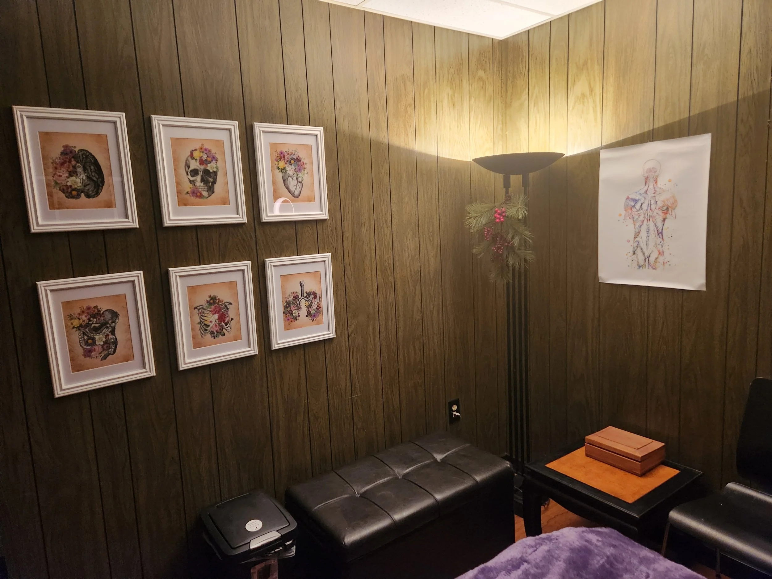 A corner of a room with wood-paneled walls, decorated with six framed pictures of skulls with floral designs, a large map or anatomical drawing, a black leather bench, a umbrella stand decorated with pine branches and red berries, a small black table with a cardboard box, and a black armchair.