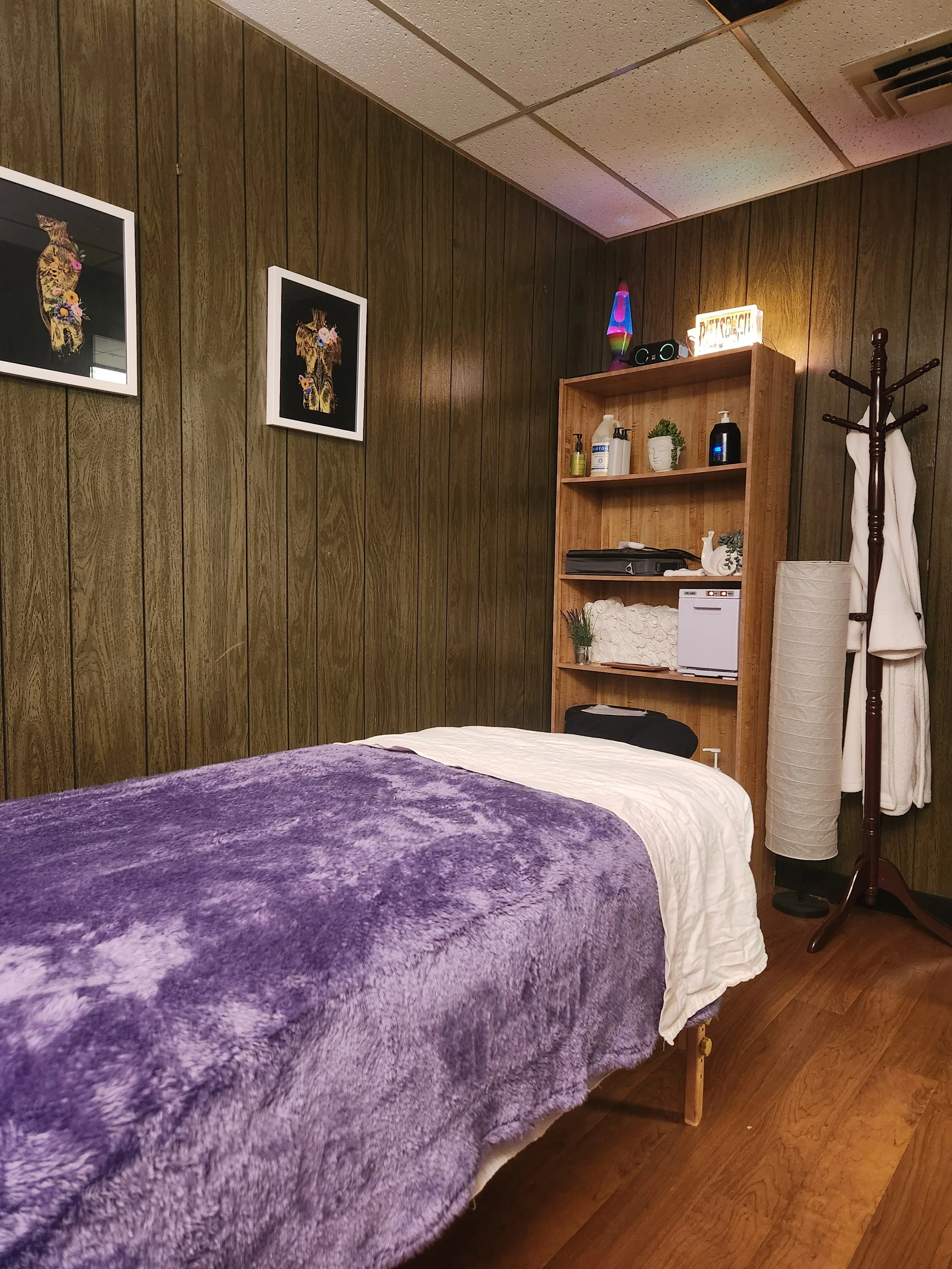 A massage table covered with a purple blanket in a room with wood-paneled walls, framed artwork of a leopard, a wooden shelving unit with various items, a coat rack with towels, and a lit lava lamp.
