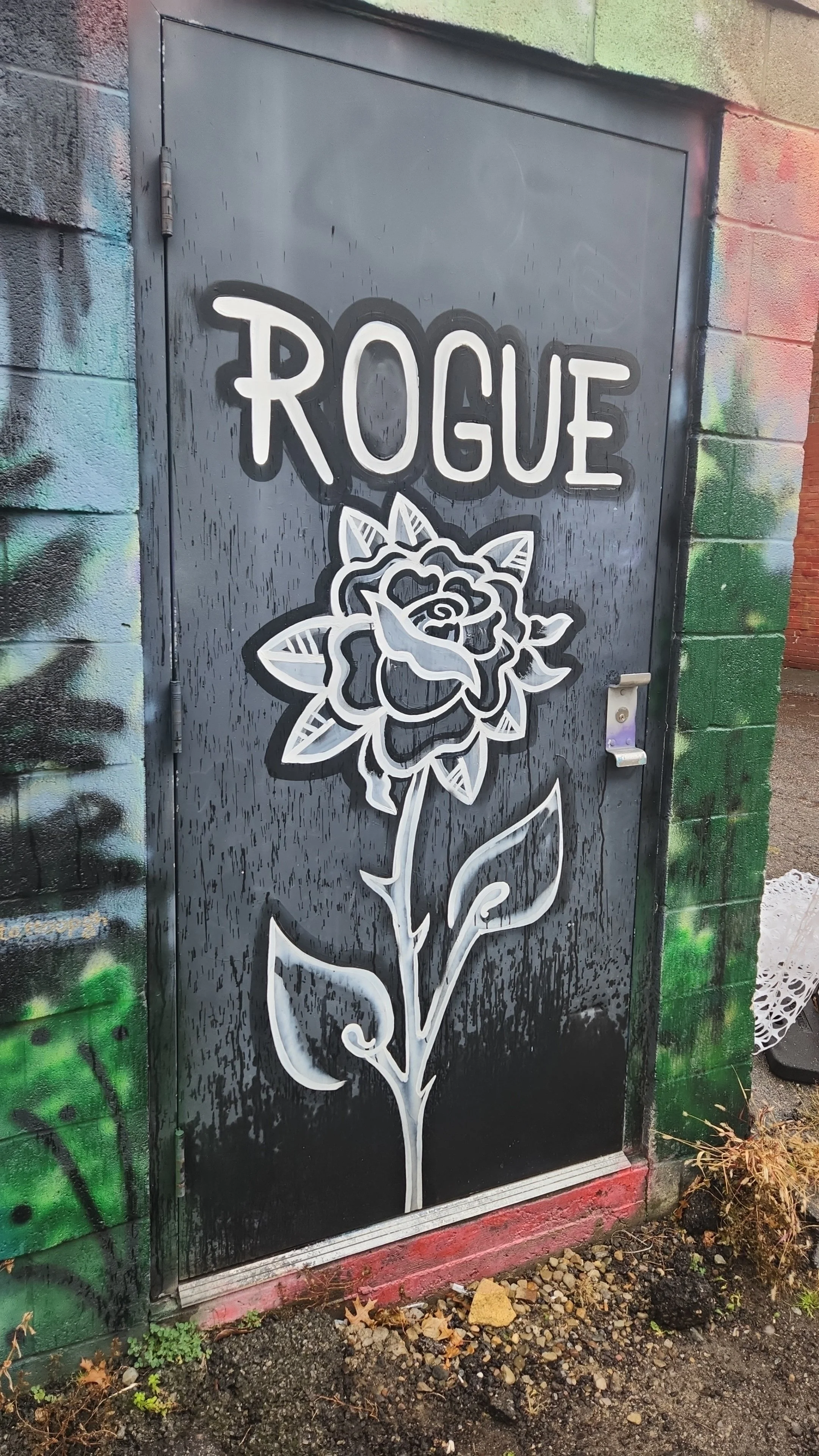 A black metal door with graffiti art that features the word "ROGUE" in white letters and a large stylized rose with detailed petals and leaves, outlined in white.