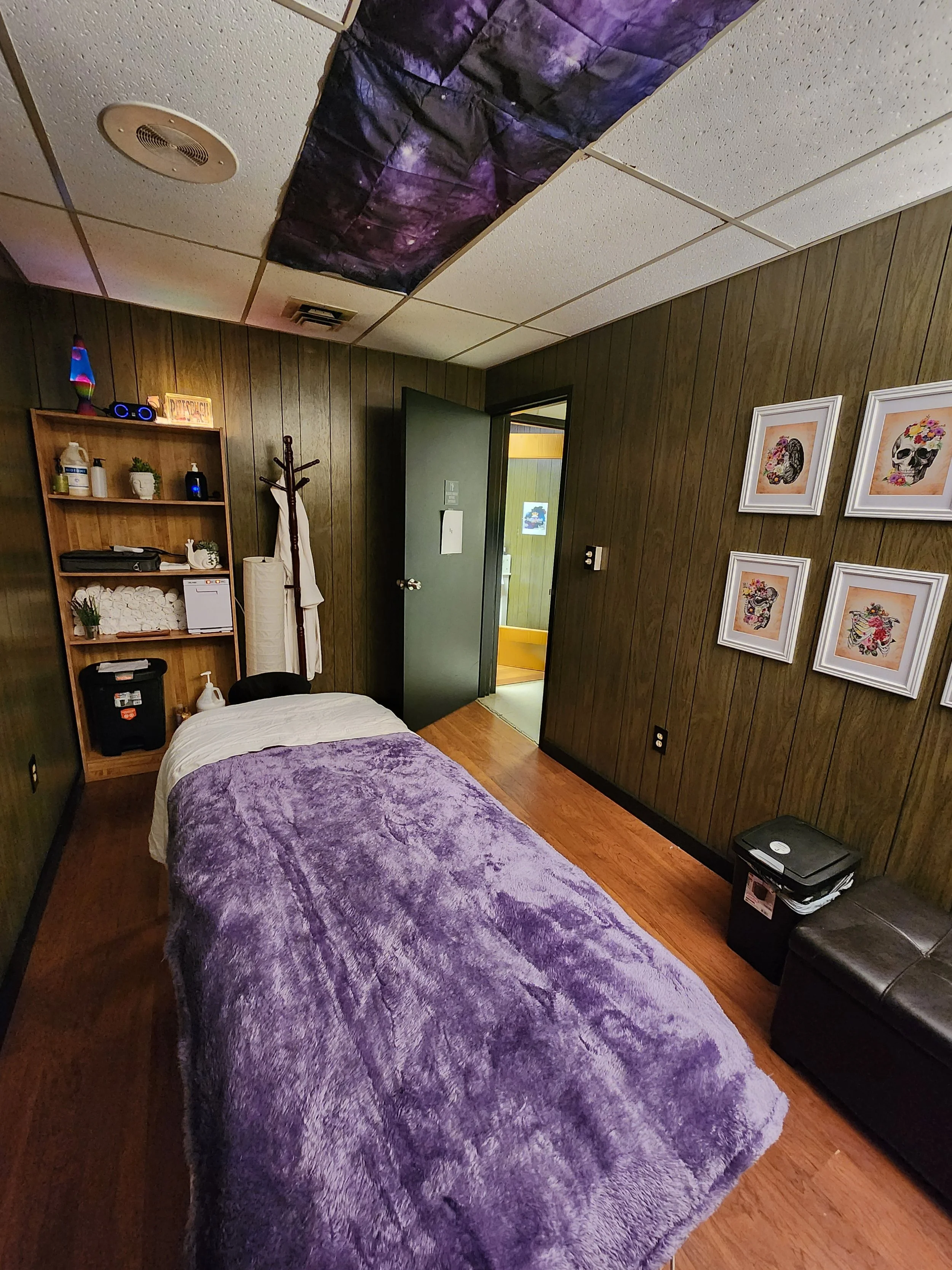 A small room with wood-paneled walls, a single bed with a purple fuzzy blanket, and framed skull art on the wall. There is a wooden shelf with various items, a coat rack, and a door leading to an adjoining room. The ceiling has purple fabric or paper decor.