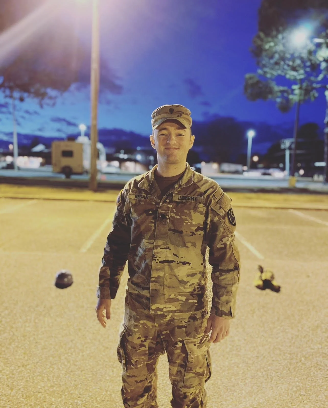 A young man in military camouflage uniform standing in a parking lot at night, smiling with eyes closed, with streetlights and trees in the background.