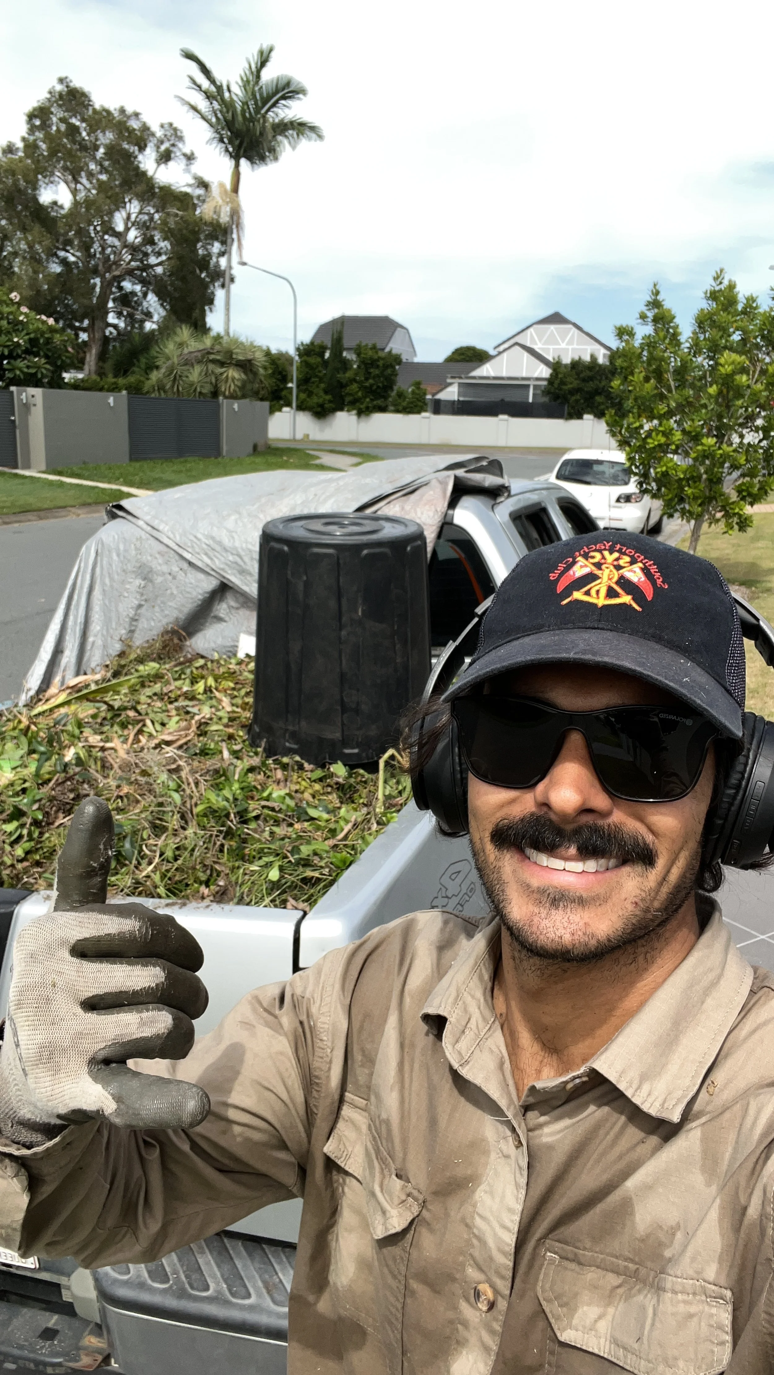 A man wearing sunglasses, a cap, and work gloves is smiling and giving a thumbs-up while taking a selfie in front of a truck loaded with yard debris, with a residential neighborhood and a palm tree in the background.