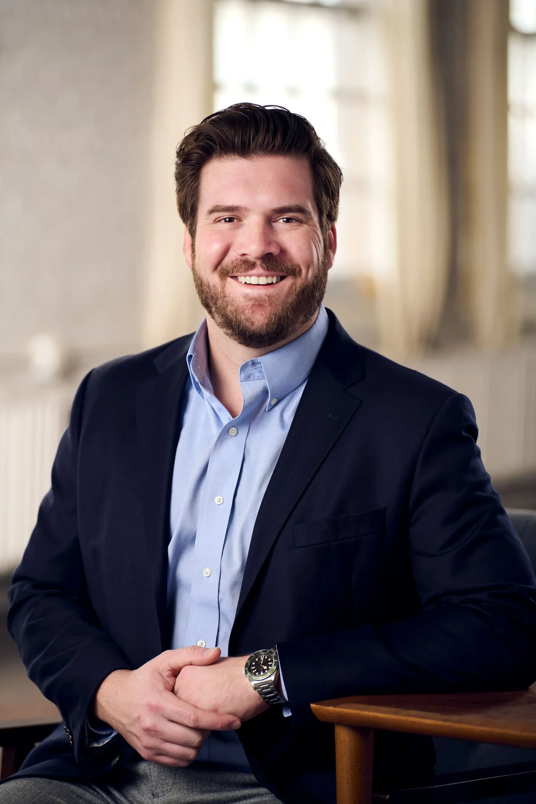 A man with a beard and brown hair, dressed in a light blue shirt and dark blue blazer, smiling and sitting in a warmly lit room with large windows and beige curtains.