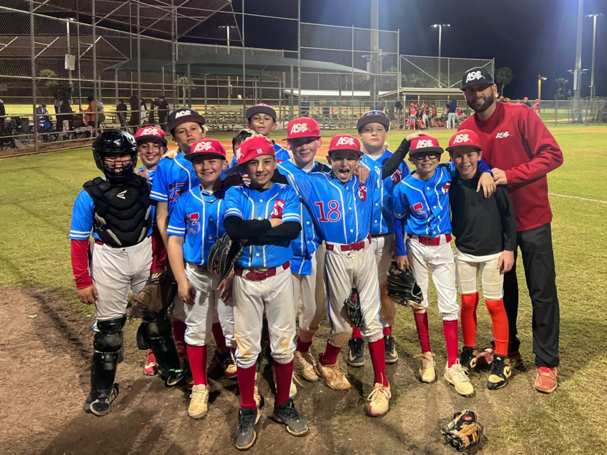 A youth baseball team posing with their coach on a baseball field at night. The players are wearing blue jerseys with red caps, white pants, and red socks. Some players are holding baseball gloves. The coach is wearing a red jacket and a black cap. The team appears to be celebrating or taking a group photo.