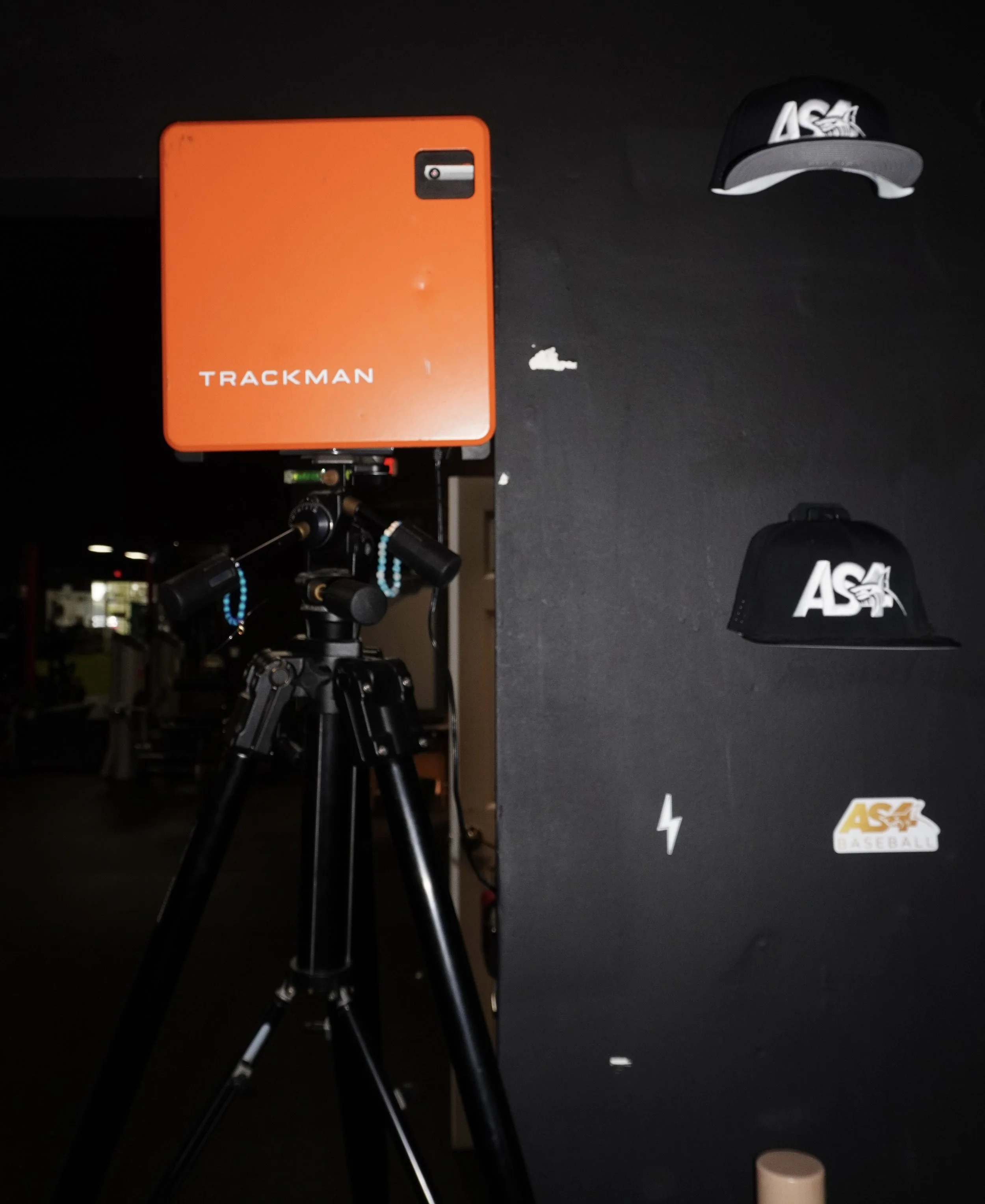 A TrackMan golf launch monitor on a tripod with hats and a lightning bolt sticker on a black wall in the background.