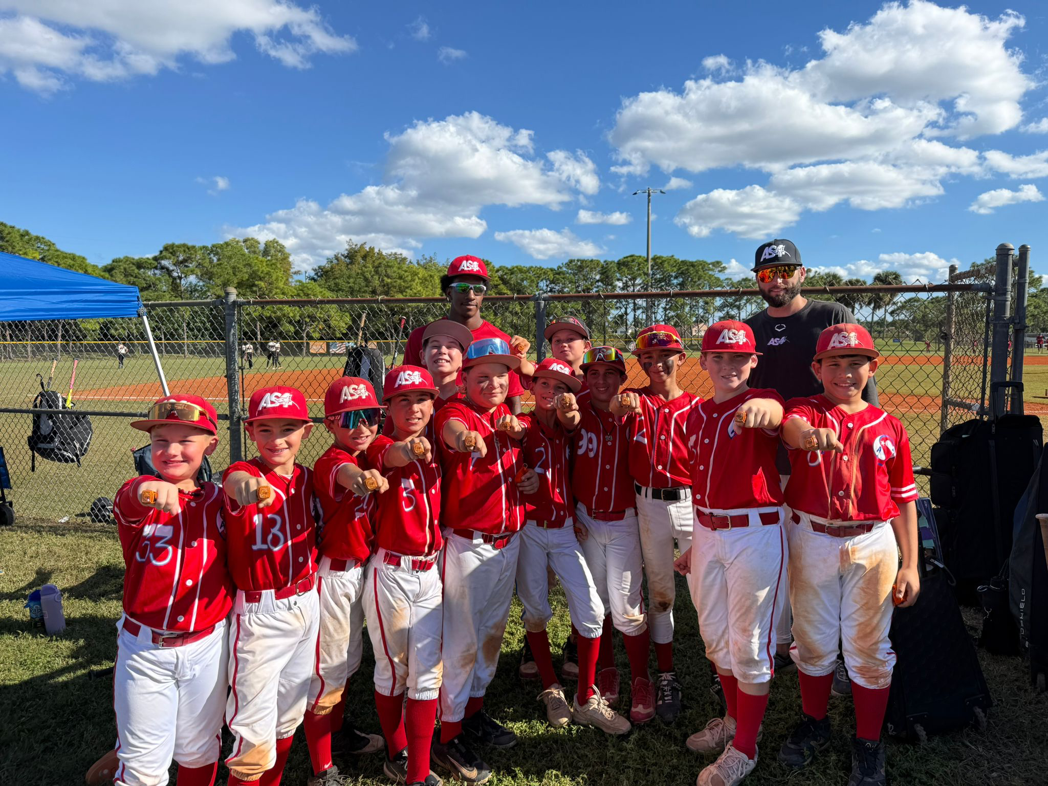 A youth baseball team dressed in red jerseys and white pants, posing together on a baseball field with their coach, all making a fist gesture, under a blue sky with some clouds.