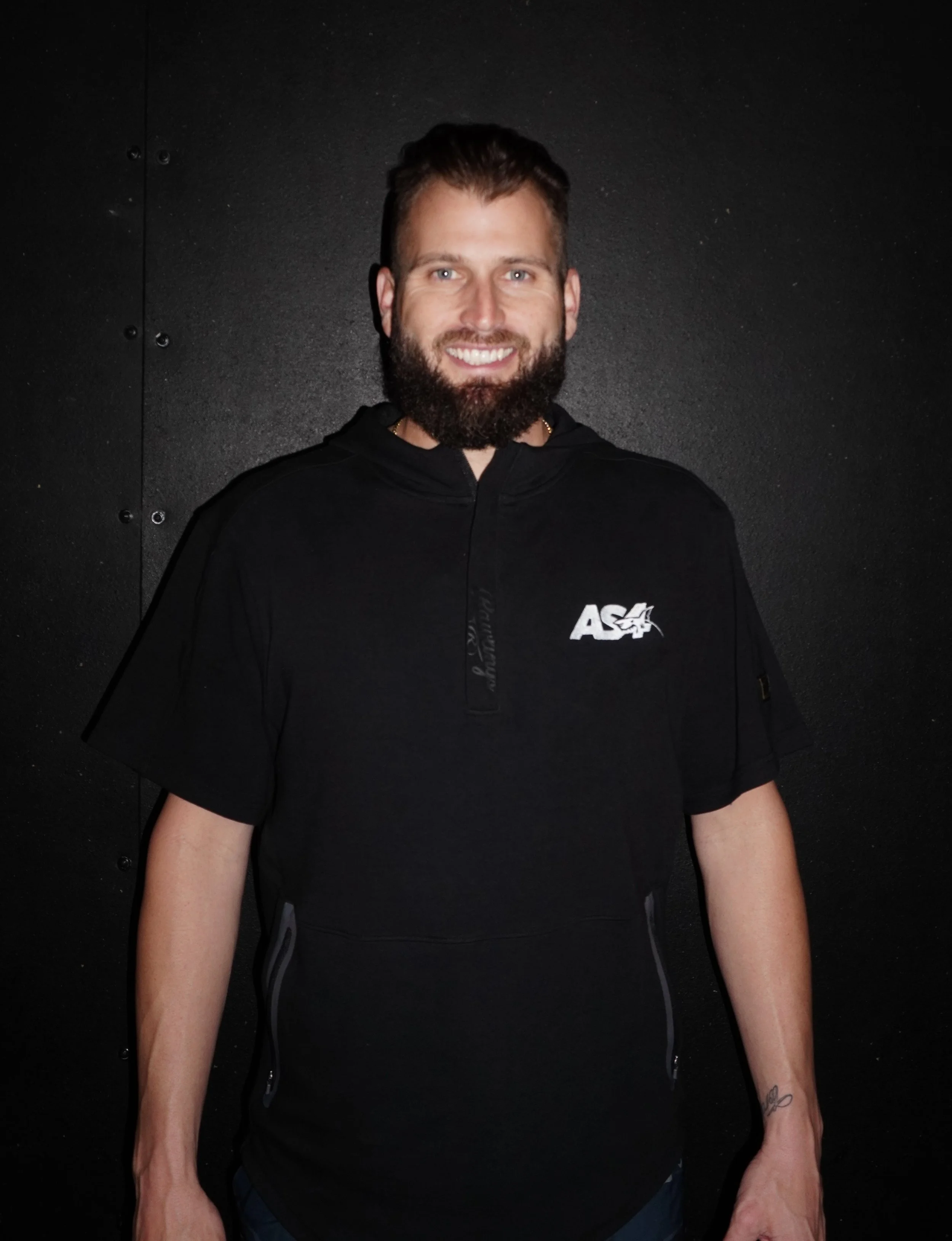 A smiling man with a beard and short hair, wearing a black NASA-themed hoodie, standing against a dark wall.