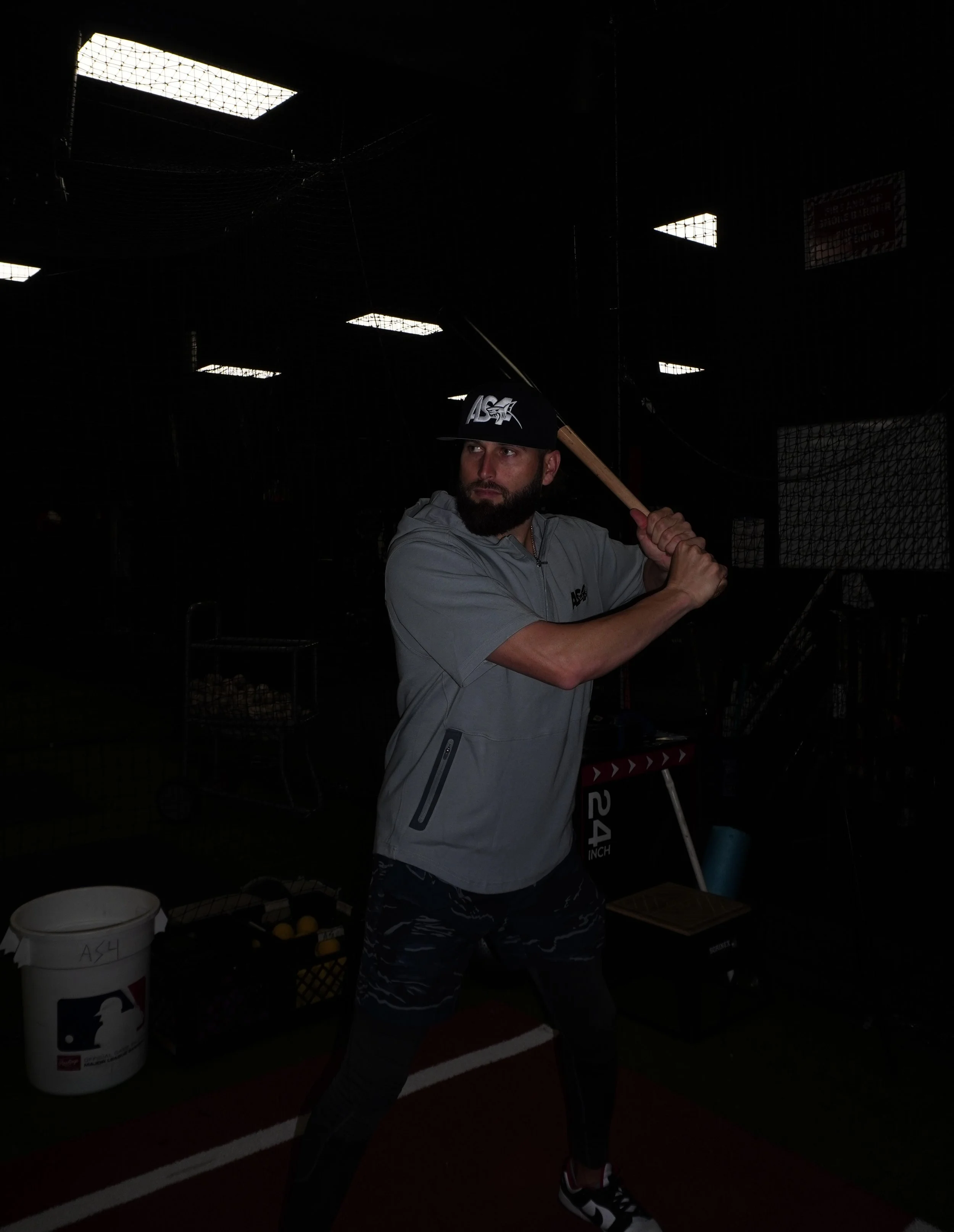 Man in gray hoodie and camouflage leggings holding a baseball bat in batting stance inside batting cage with bright ceiling lights.