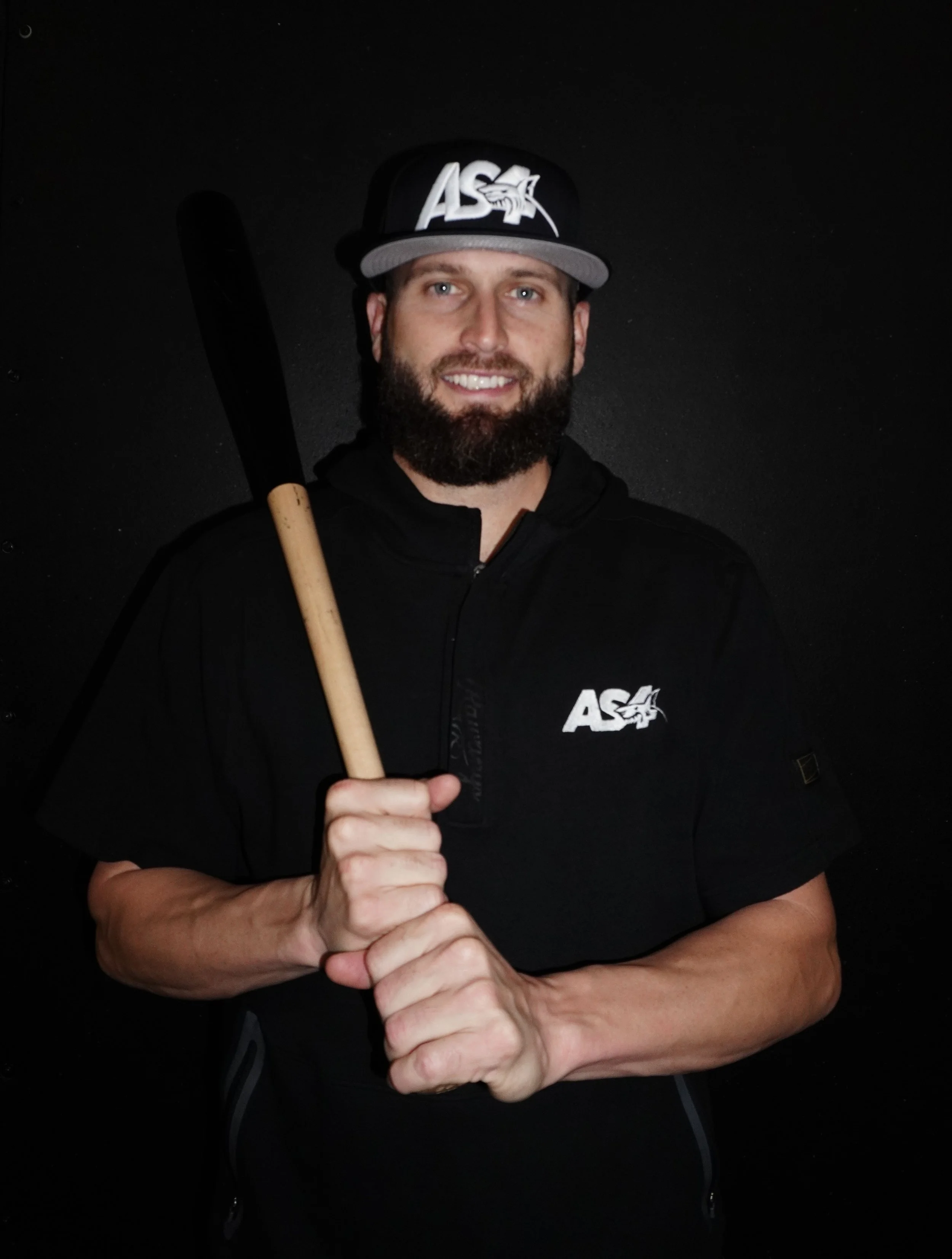 A man with a beard smiling, holding a baseball bat, wearing a black cap and black hoodie with a logo, against a black background.