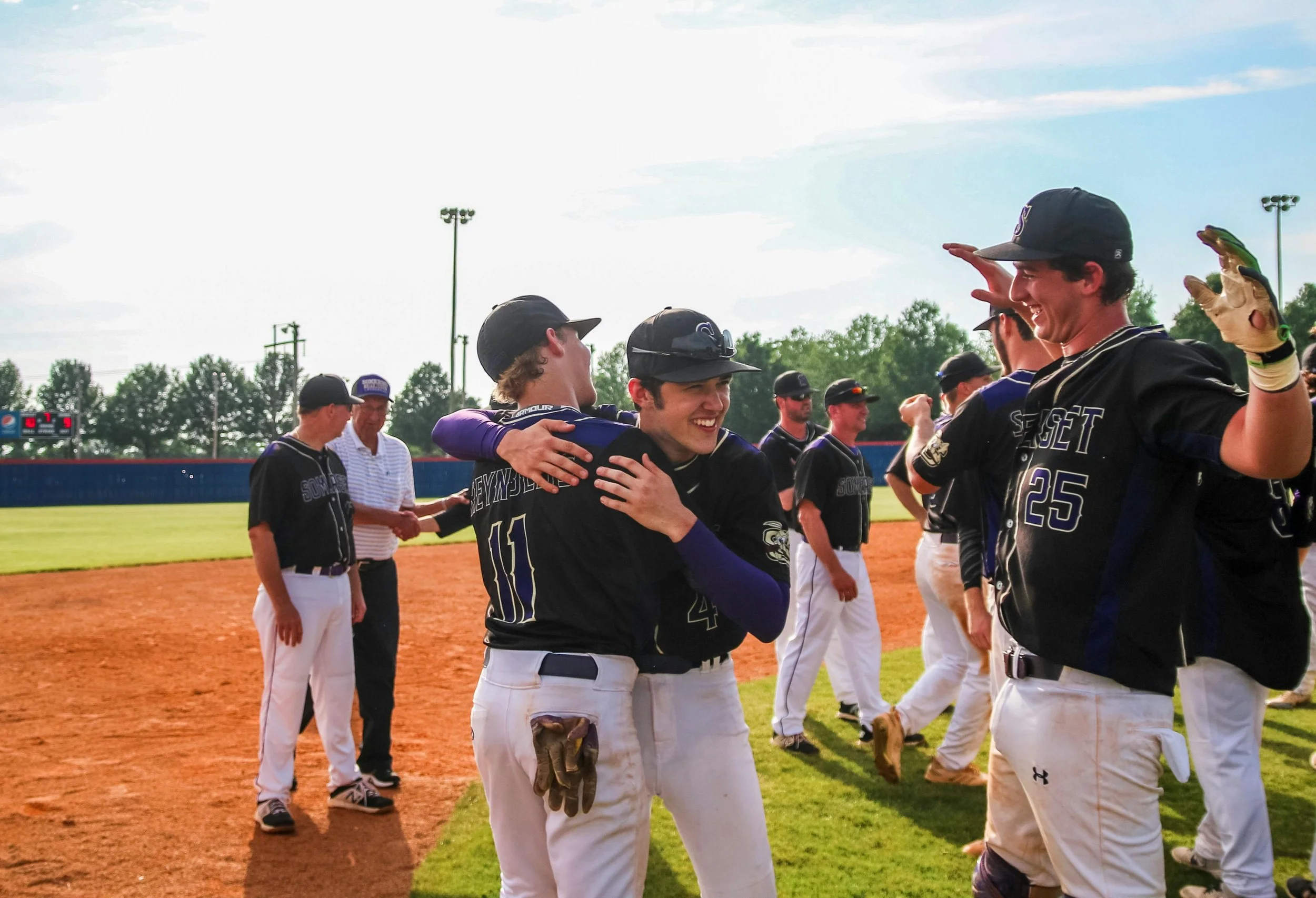 Baseball players celebrating on the field with smiles and hugs, wearing black and white uniforms, with a baseball diamond and green trees in the background.