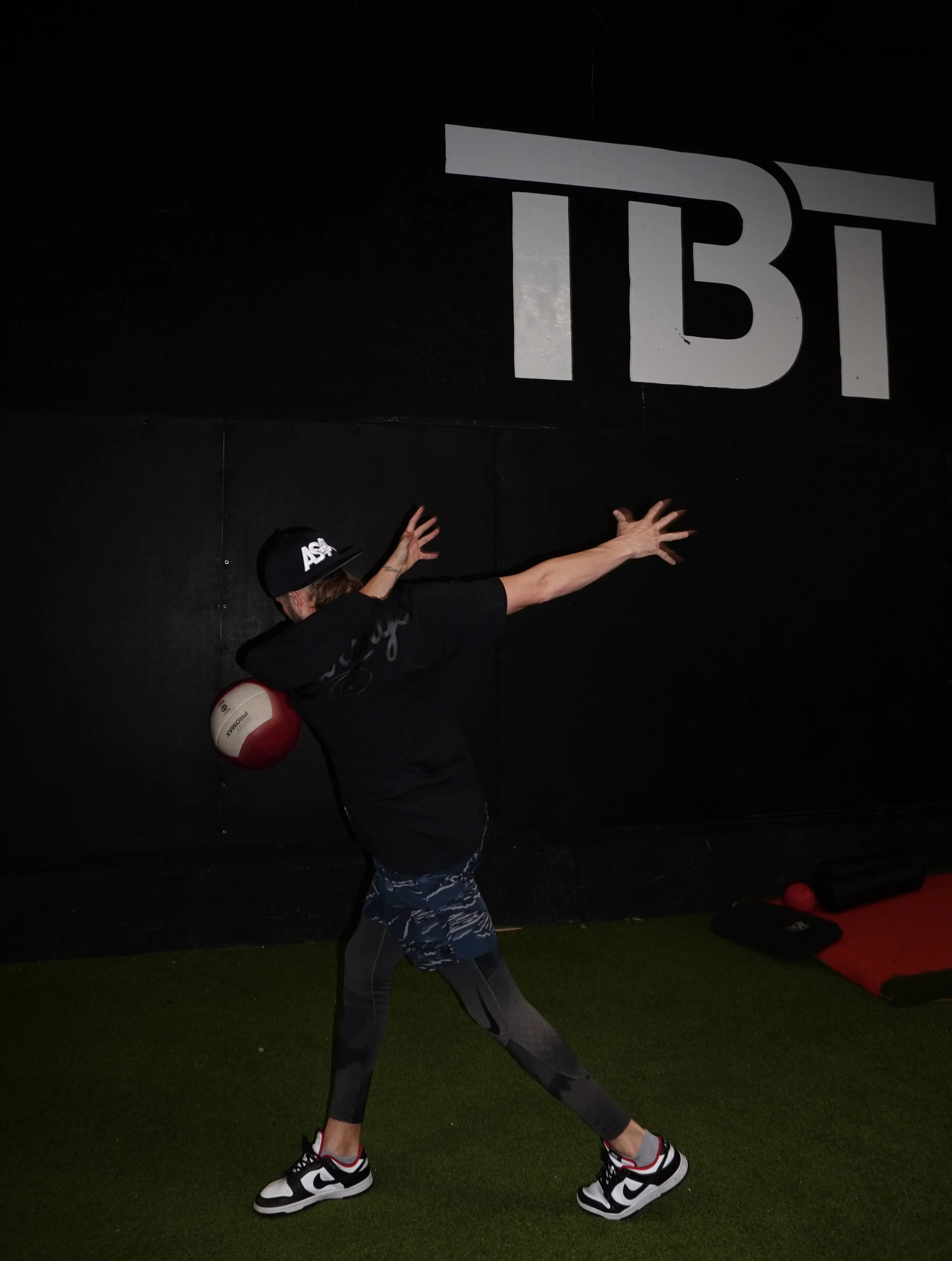 A person wearing a black shirt, camo shorts, sunglasses, a black cap, and sneakers is performing a stretching exercise on a green turf floor inside a gym. The person is holding a medicine ball against their head with one arm while stretching the other arm outward. A large sign with the letters 'TBT' is visible on the black wall behind them.