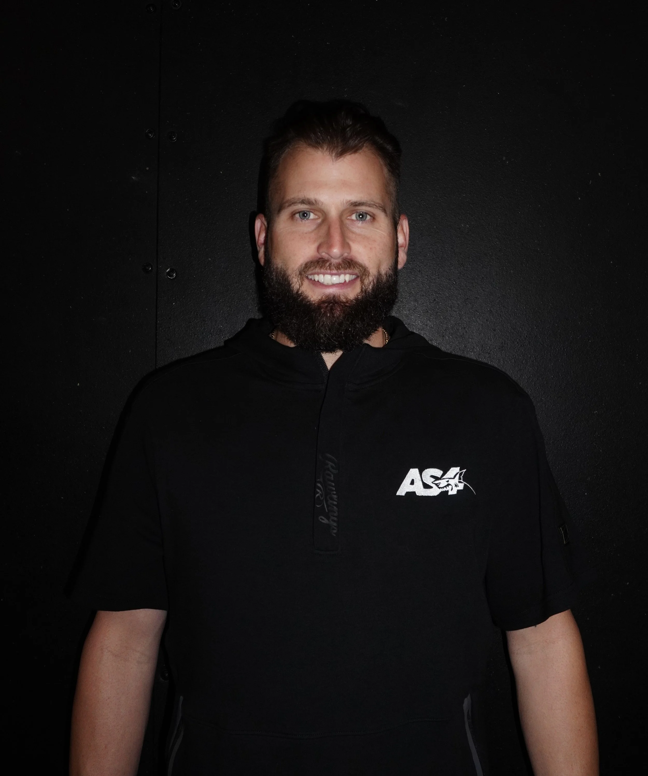 A smiling man with a beard and short hair, wearing a black hoodie with 'AS4' and a shark logo, standing against a black wall.
