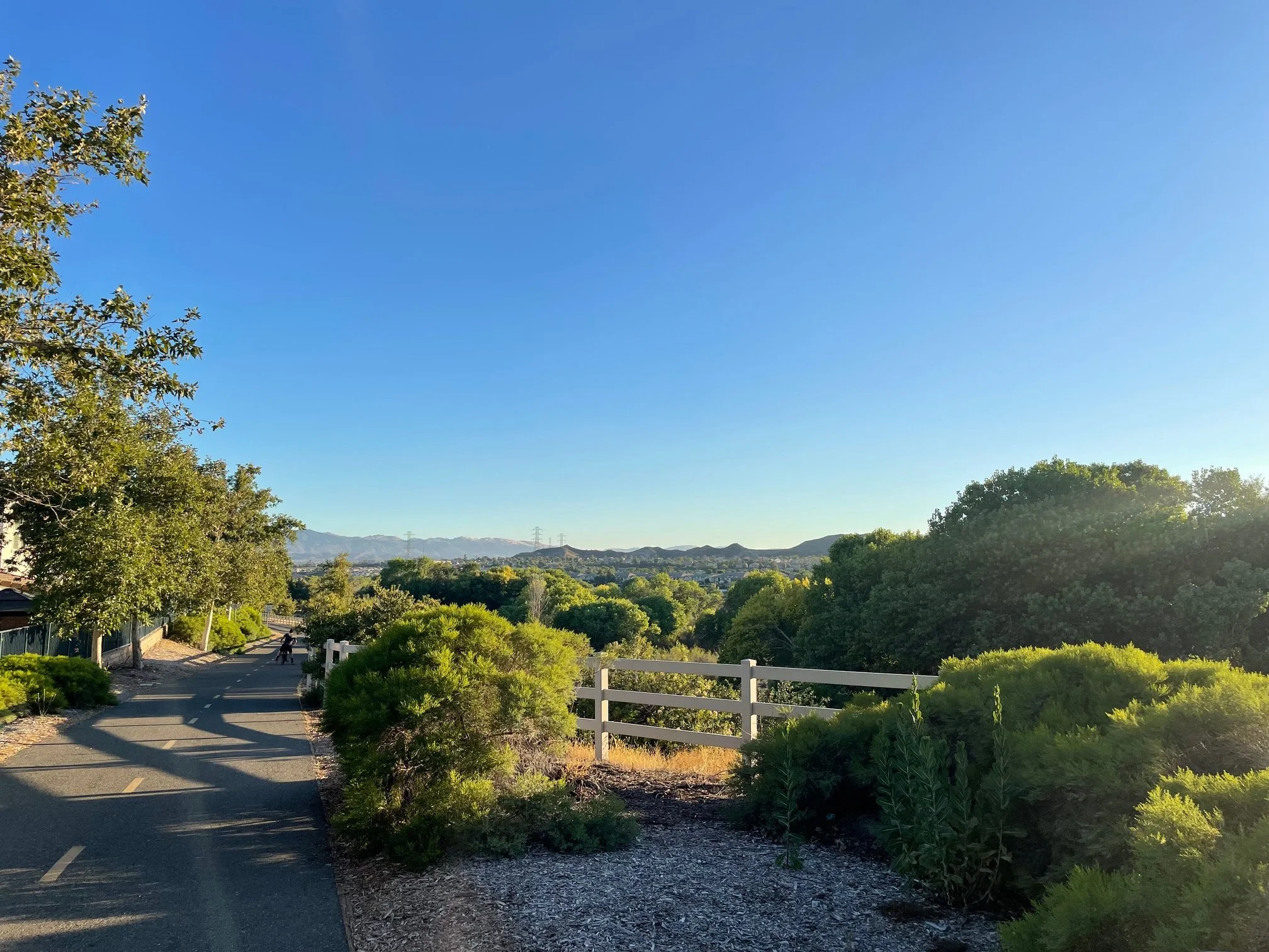 A scenic view of a clear blue sky over a green park with trees and shrubs. A paved walking trail runs through the park, with a person walking a dog in the distance. White fencing lines part of the trail, and rolling hills are visible in the background.