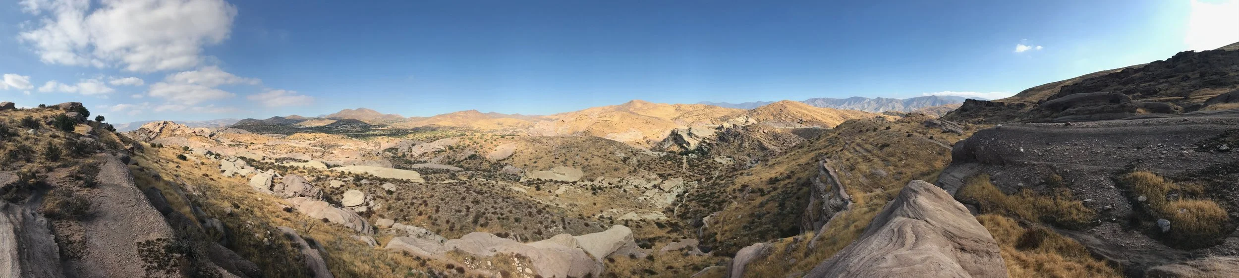 Panoramic view of a desert landscape with rocky hills, sparse vegetation, and distant mountains under a blue sky with a few clouds.