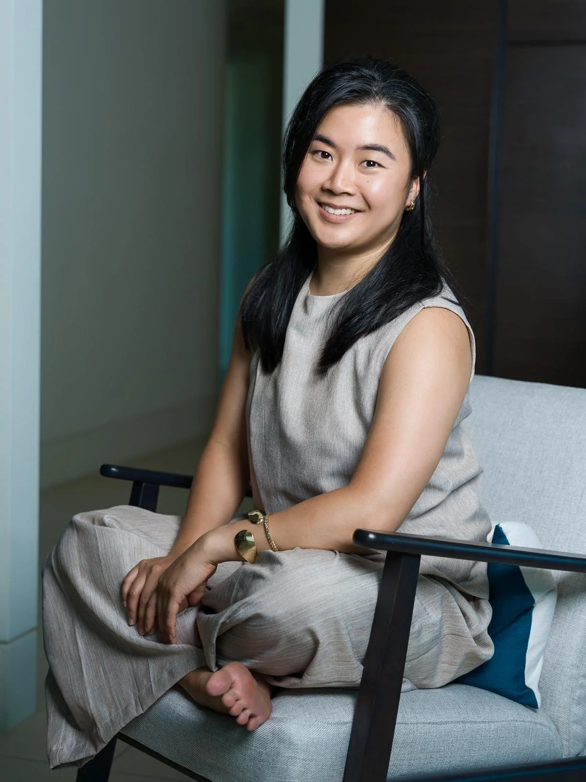 A young woman with black hair, smiling, sitting barefoot in a modern room on a beige chair with black armrests, wearing a beige sleeveless dress and gold jewelry.
