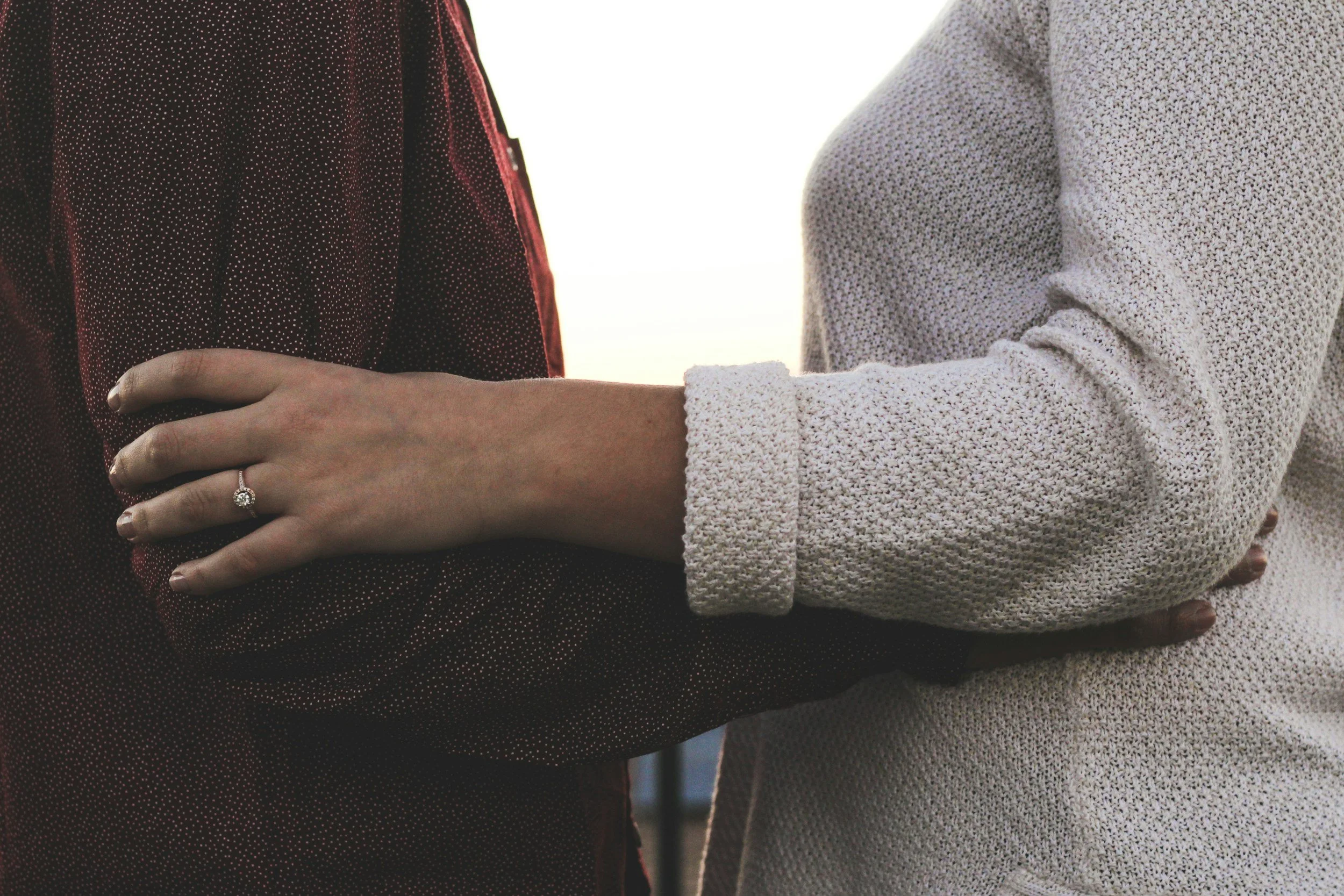 Two women are holding hands, one with a ring on her finger. One woman is wearing a dark red and black polka dot dress, and the other is wearing a light-colored textured sweater. The focus is on their hands and the ring.