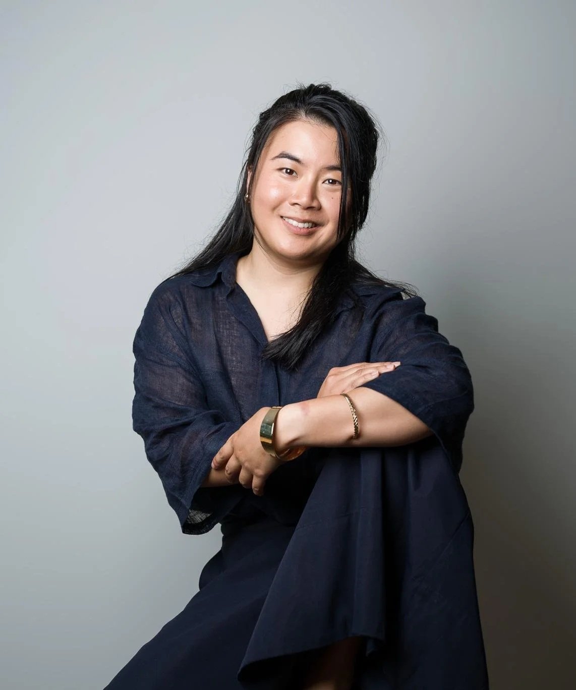 Portrait of a woman with black hair wearing a dark blue shirt, smiling, sitting with arms crossed in front of a plain gray background.