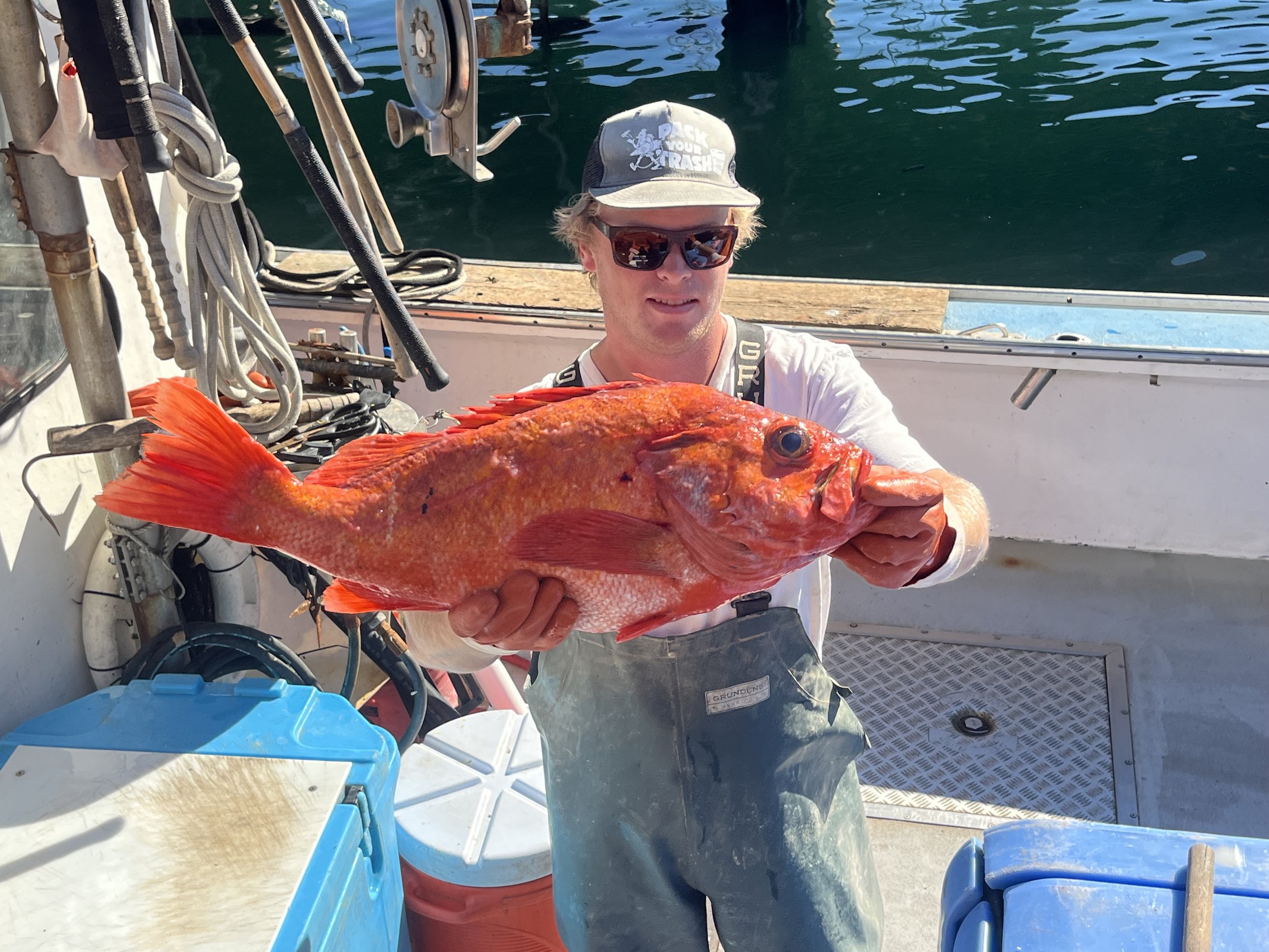 A young man on a boat holding a large red fish, wearing sunglasses, a hat, and rubber overalls.