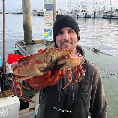 Man in a black beanie holding a large crab at a marina with boats docked in the background.