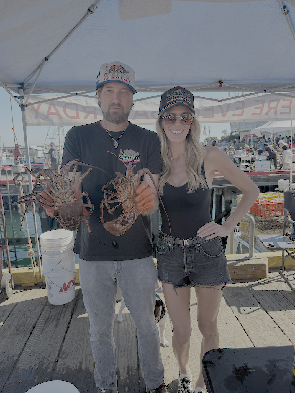 Two people at a seafood market or dock holding lobsters, wearing casual clothes and hats, with a tent and water in the background.