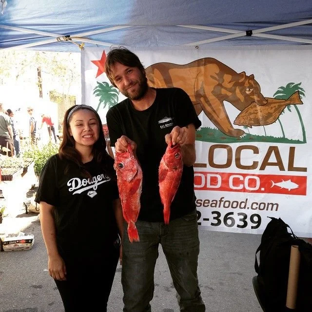 A man and woman smiling at a seafood market stall, holding two large red fish. The stall has a banner with a bear and palm trees, advertising Local Food Co.