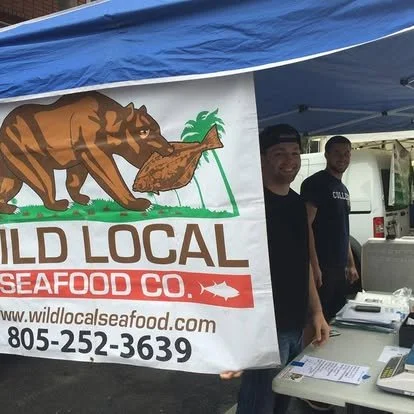 Food stall for Wild Local Seafood Co. with a banner showing a bear carrying seafood, two men behind the counter, under a blue canopy.