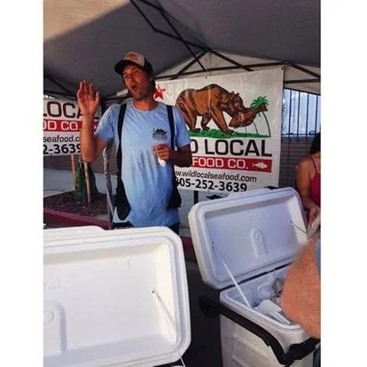 Man in a blue shirt and cap at a seafood booth, waving, with a banner displaying a bear and a pig in the background.