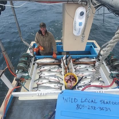 A man on a fishing boat displaying a freshly caught fish, with multiple fish laid out on the boat's deck and a cooler filled with fish.
