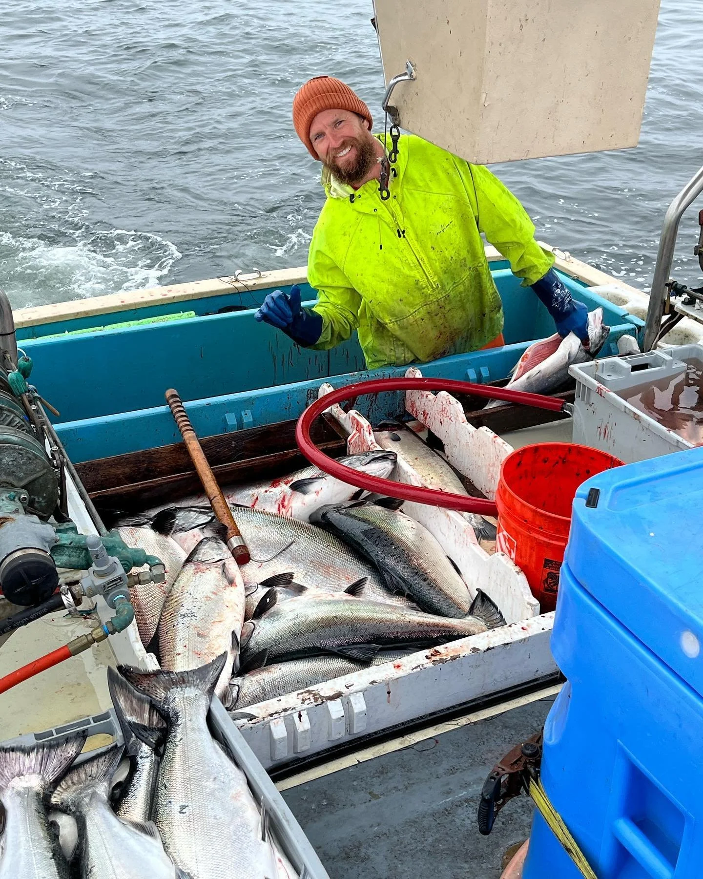 A man in a yellow jacket and orange beanie on a boat, smiling with fish caught from fishing, with a cooler full of fish.