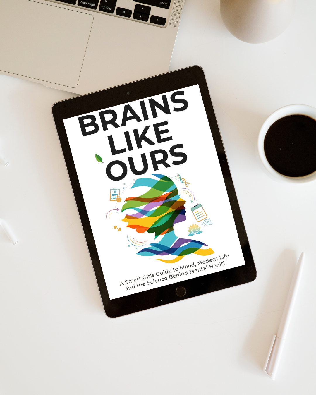 A tablet with the book cover titled "Brains Like Ours: A Smart Girls Guide to Mood, Modern Life and the Science Behind Mental Health" on a white desk. Nearby are a laptop, a cup of coffee, a white pen, and a gray vase.