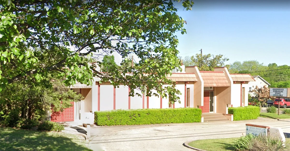 A small building with white walls, red accents, and a red door, surrounded by green trees and bushes, with a parking lot in front and a blue sky overhead.