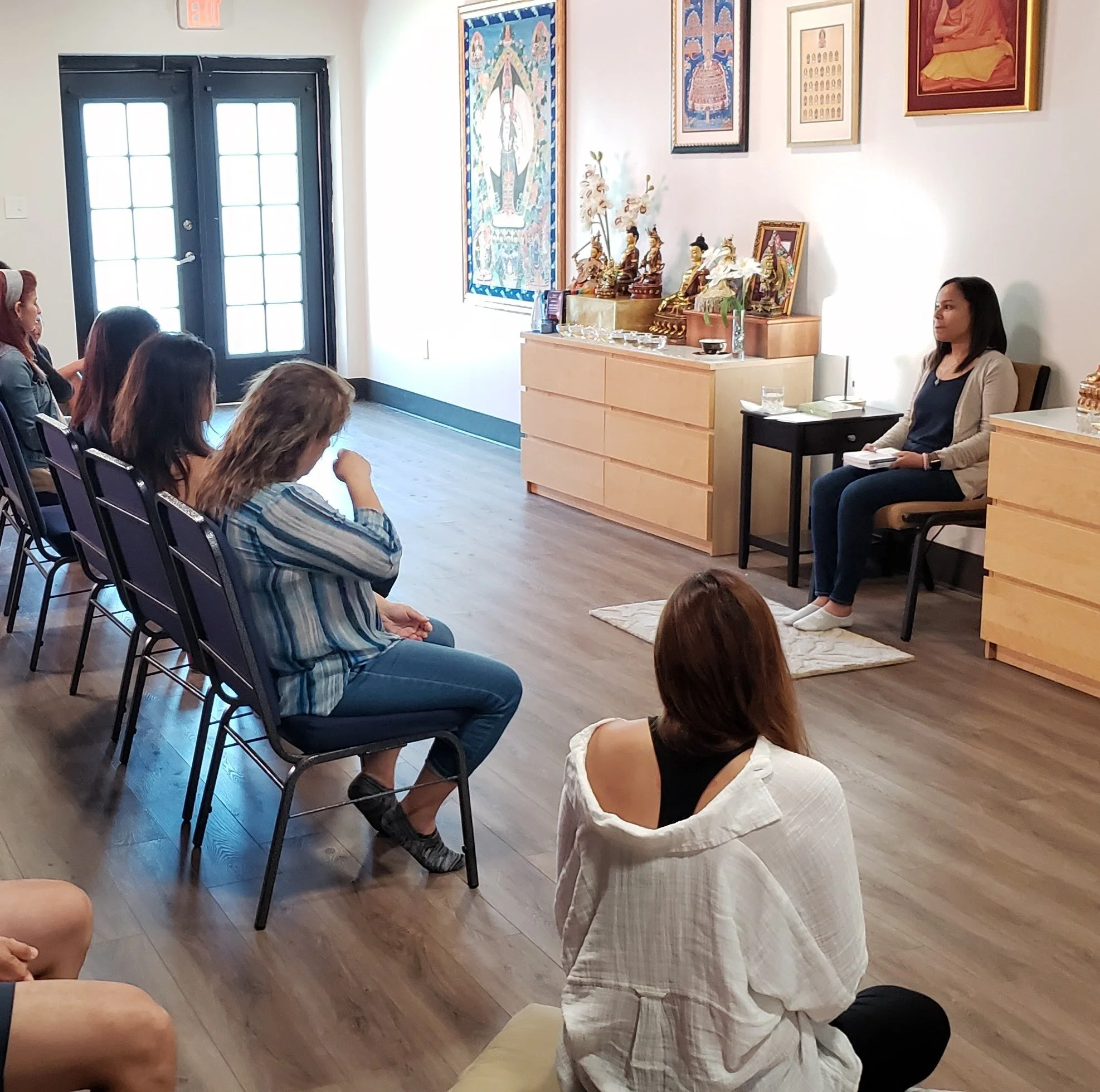 A woman sits on a chair in front of a small group of women in a room with Buddhist artwork and statues, possibly leading a meditation or spiritual session.